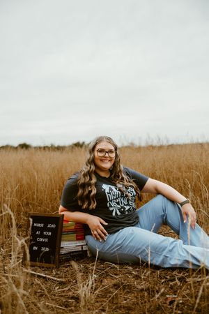 Nursing graduate sits in golden field holding books with letter board sign reading Nurse Jojo At Your Cervix RN 2025.