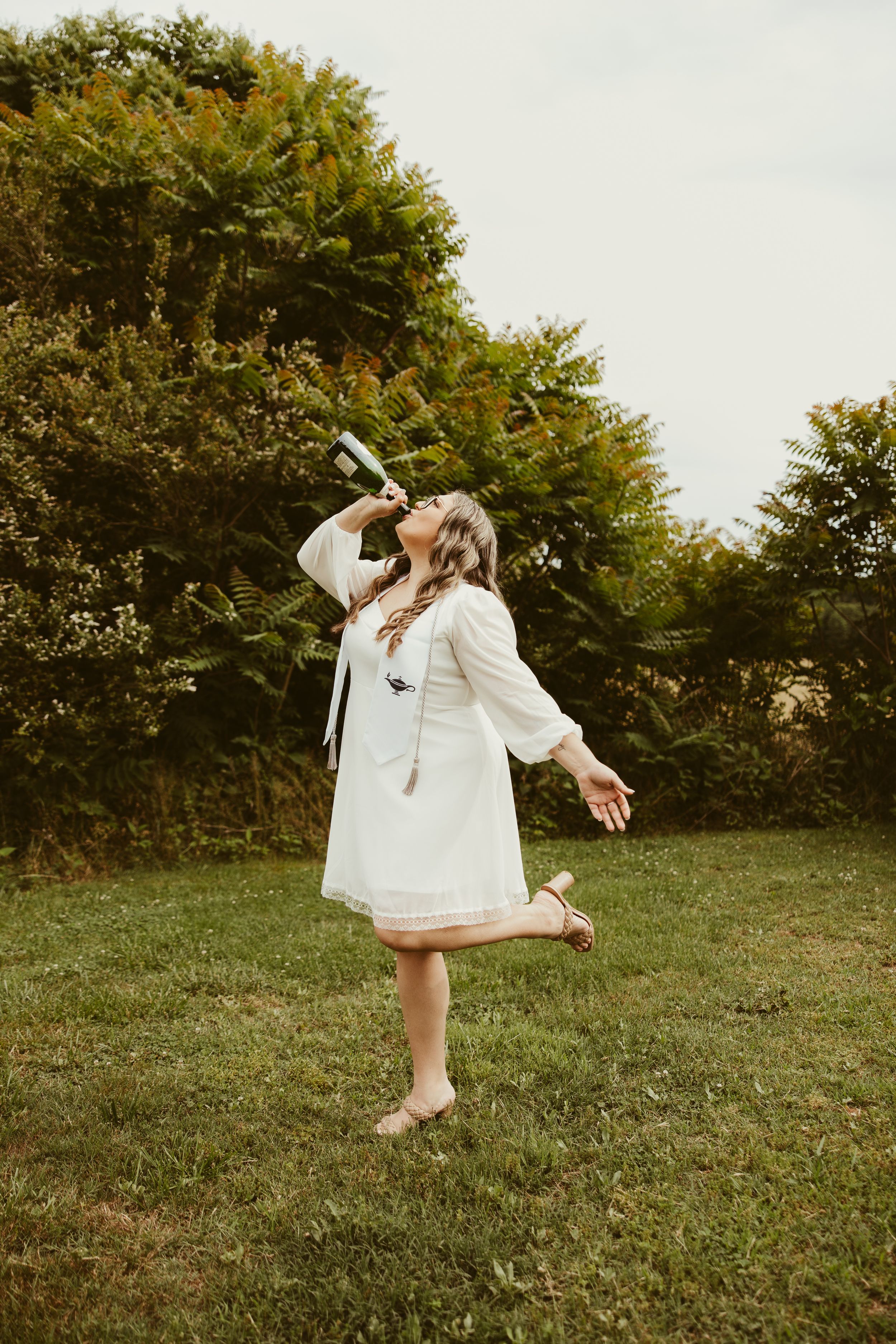 Woman in white dress celebrates outdoors drinking champagne barefoot in green garden setting.