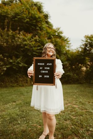 Nursing graduate holds letter board sign reading Nurse Jojo At Your Cervix RN 2025 in garden.