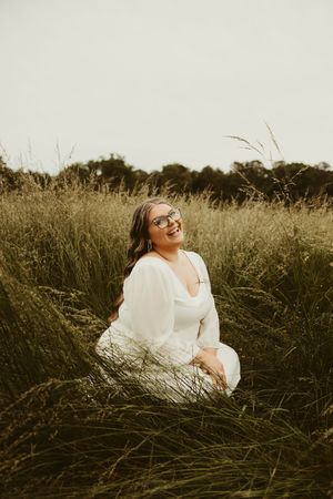 Woman in white dress smiles joyfully while sitting in tall green grass field on overcast day.