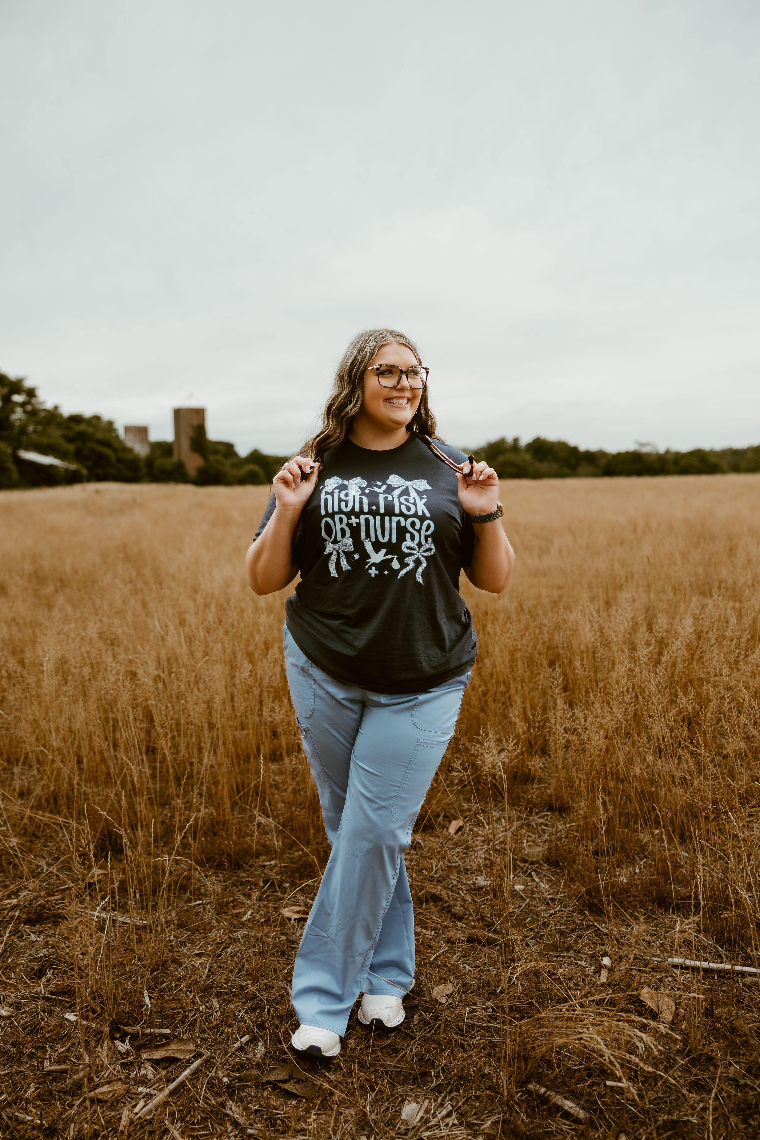 Smiling woman in graphic tee and flare jeans standing in golden wheat field on overcast day.