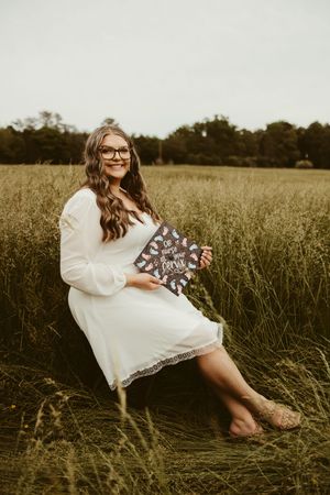 Smiling nursing graduate in white dress holds decorated cap sitting in lush green field.