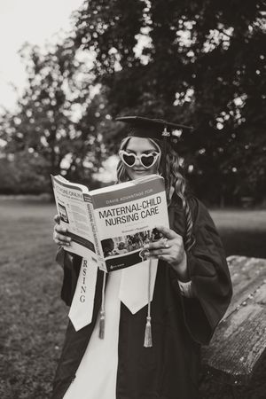 Nursing graduate in cap and gown reads Maternal-Child Nursing Care textbook outdoors in black and white photo.