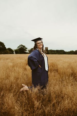 Smiling nursing graduate in navy cap and gown poses in golden wheat field on cloudy day.
