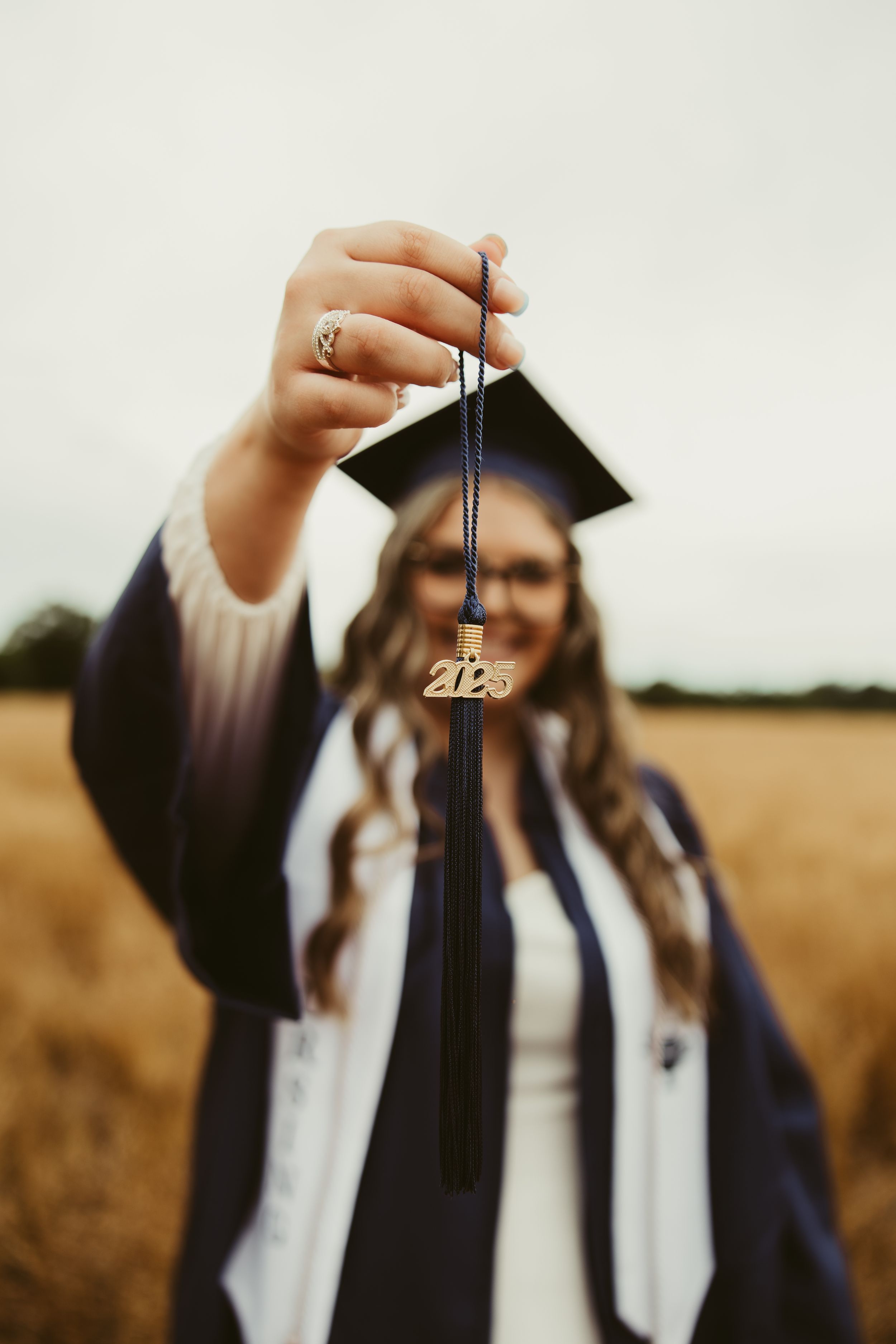 Nursing graduate holds up 2025 graduation tassel attached to cap in golden field.