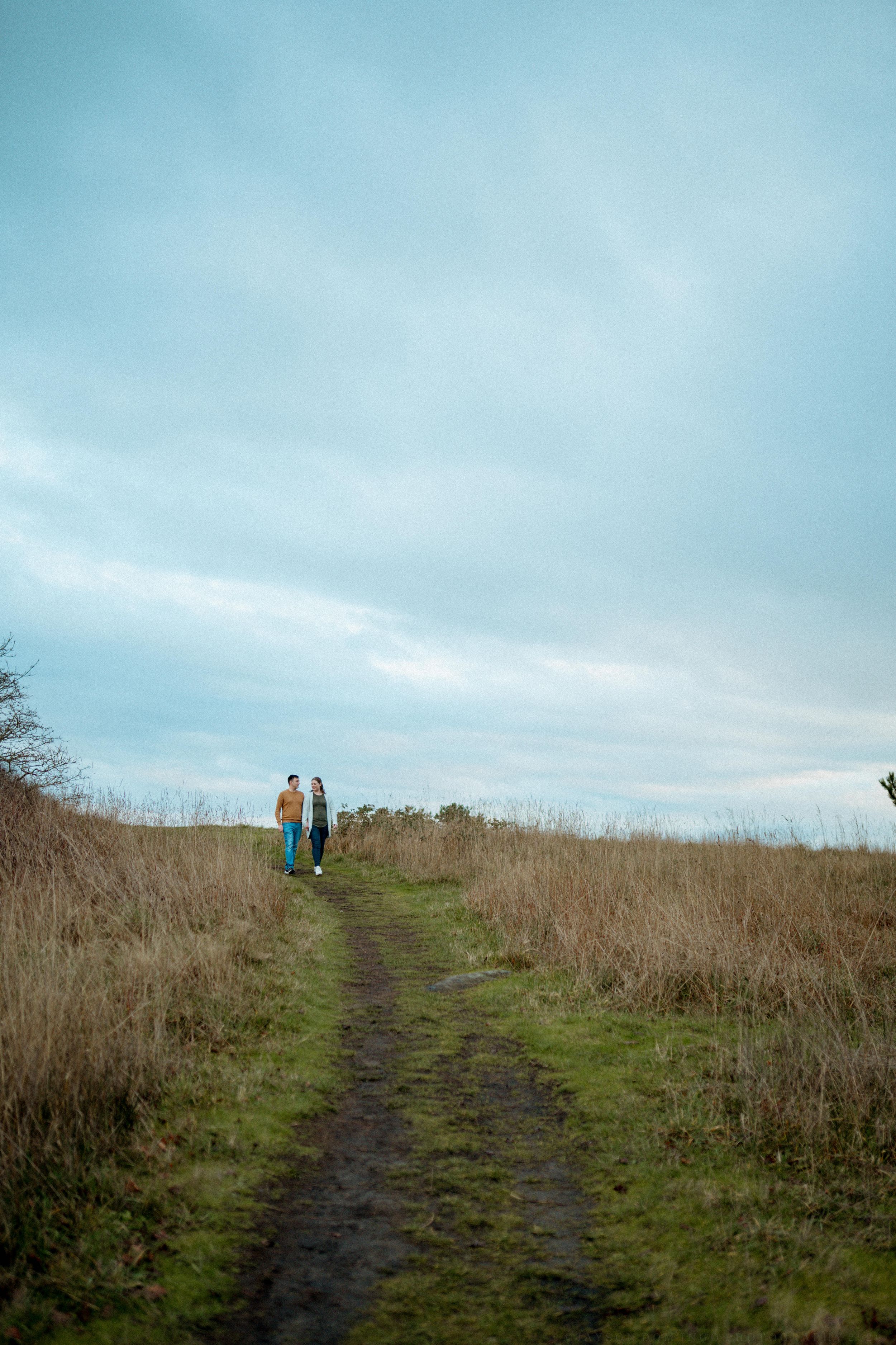 Two people walk along a grassy hilltop trail against a bright blue sky on a sunny day.