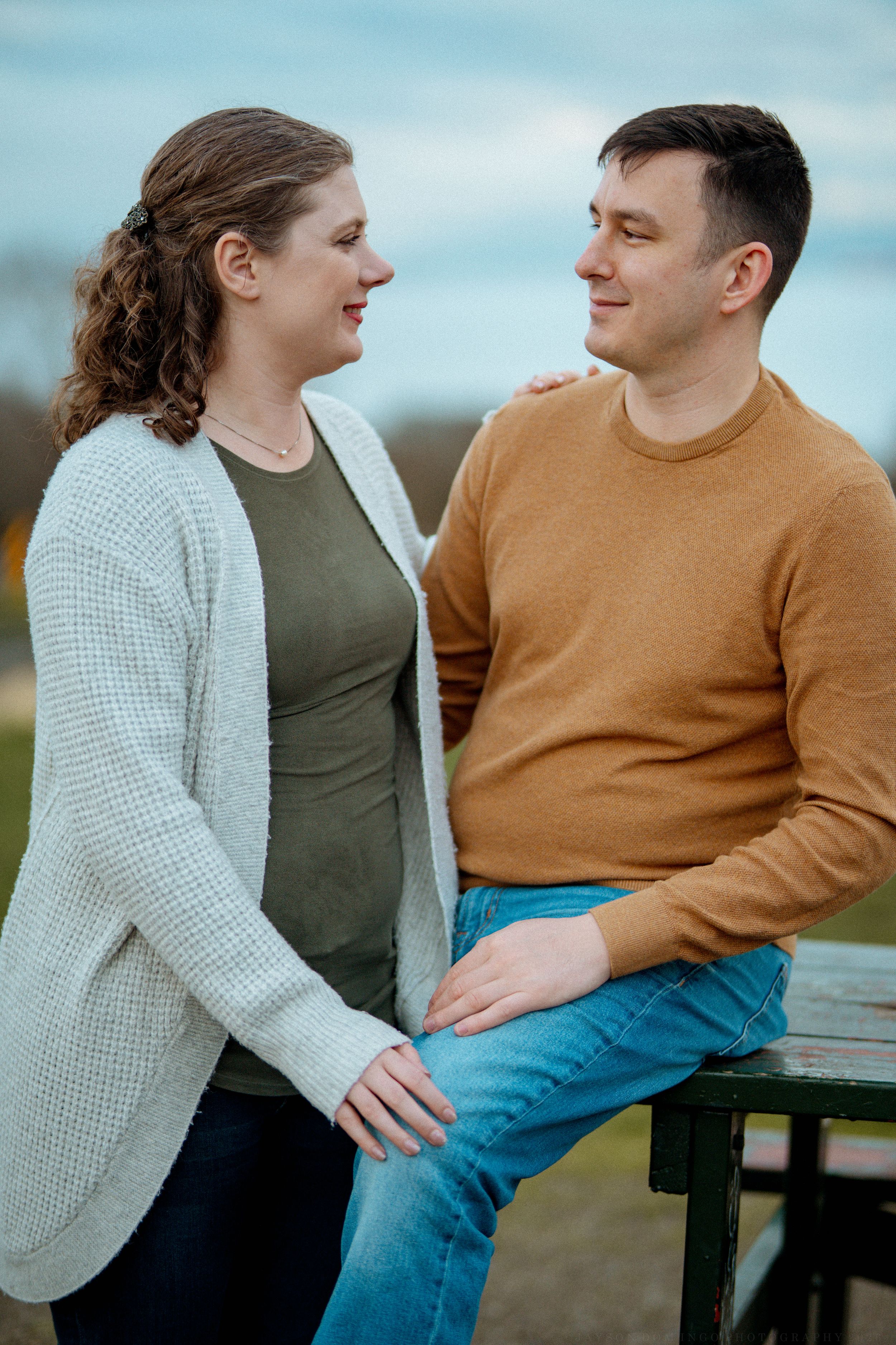 A couple sits together on a wooden bench outdoors sharing candid moments and laughter in casual autumn attire.