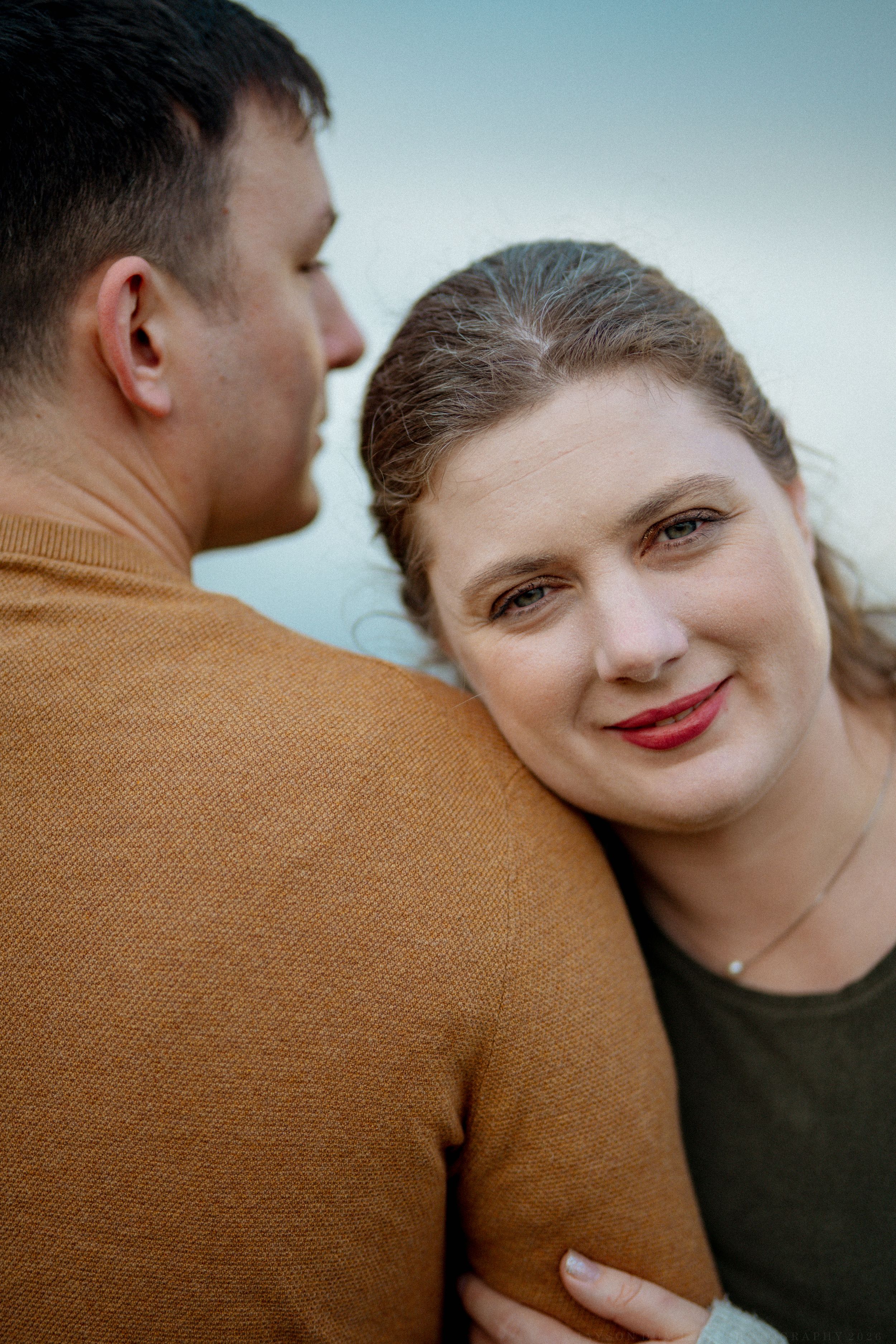 A couple shares a tender moment outdoors with one person wearing a brown sweater against a blue sky background.