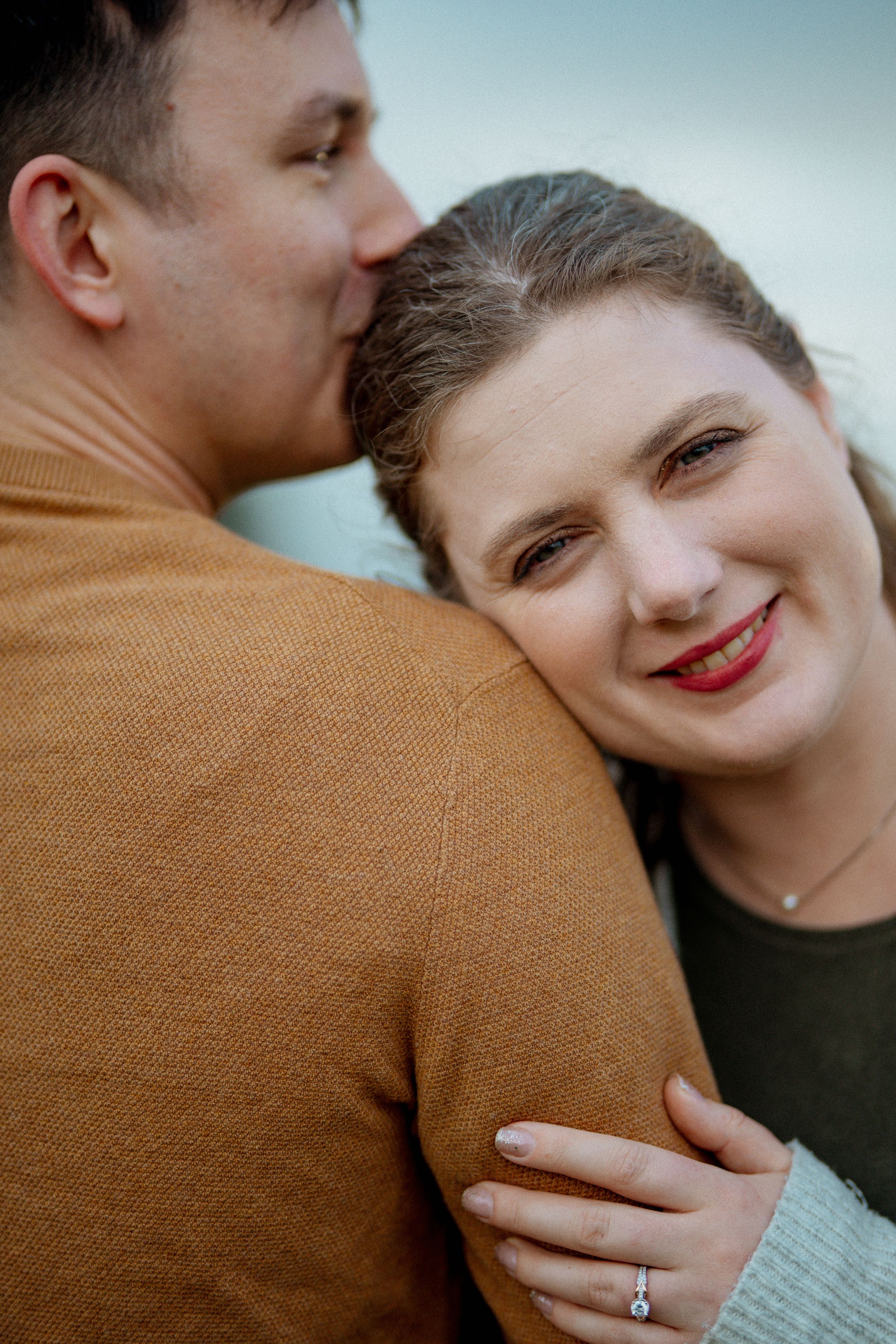 Close-up engagement photos showing tender moments and a sparkling ring against a brown sweater background.