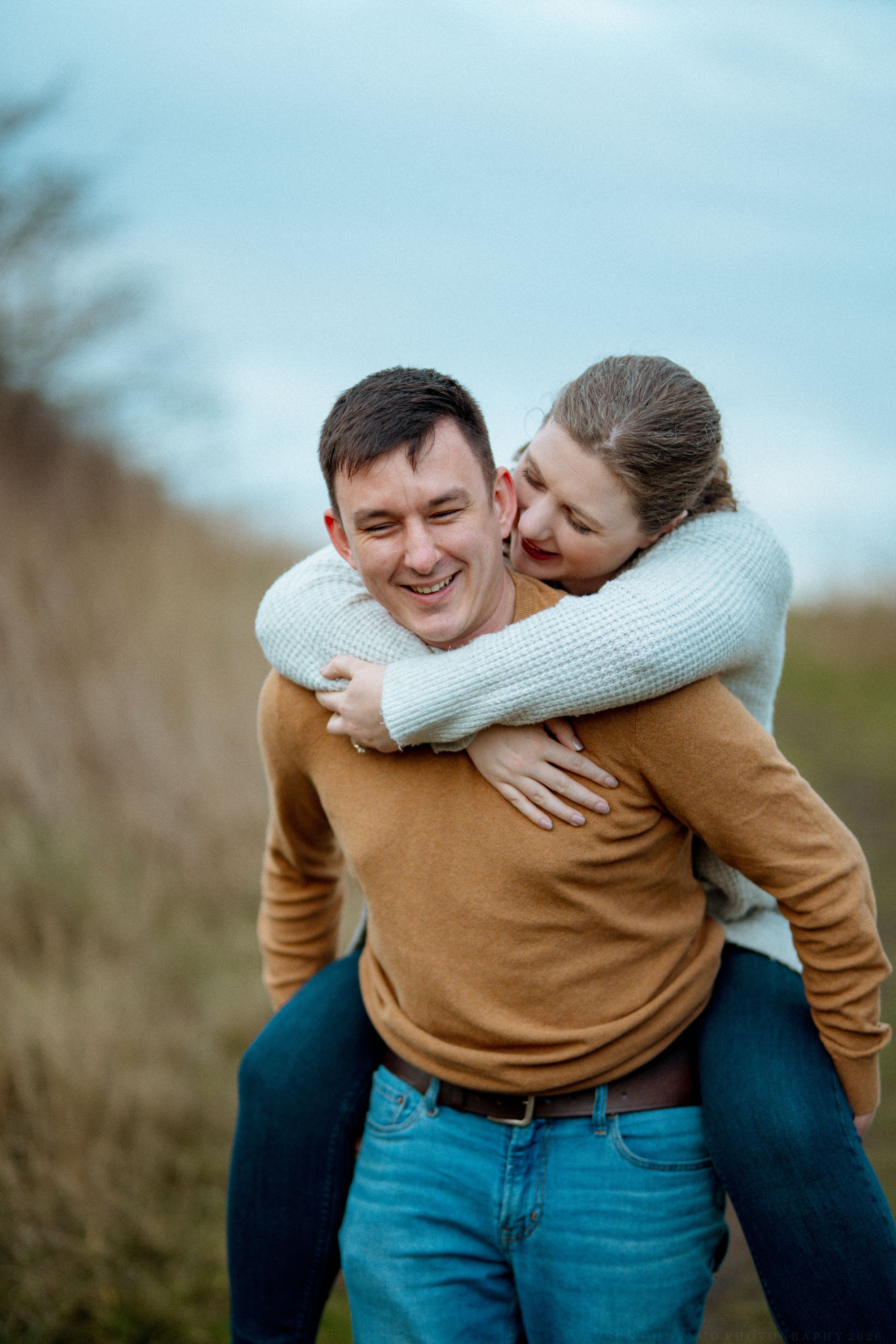 A playful couple shares a joyful moment outdoors, with one person giving a piggyback ride in casual autumn attire.