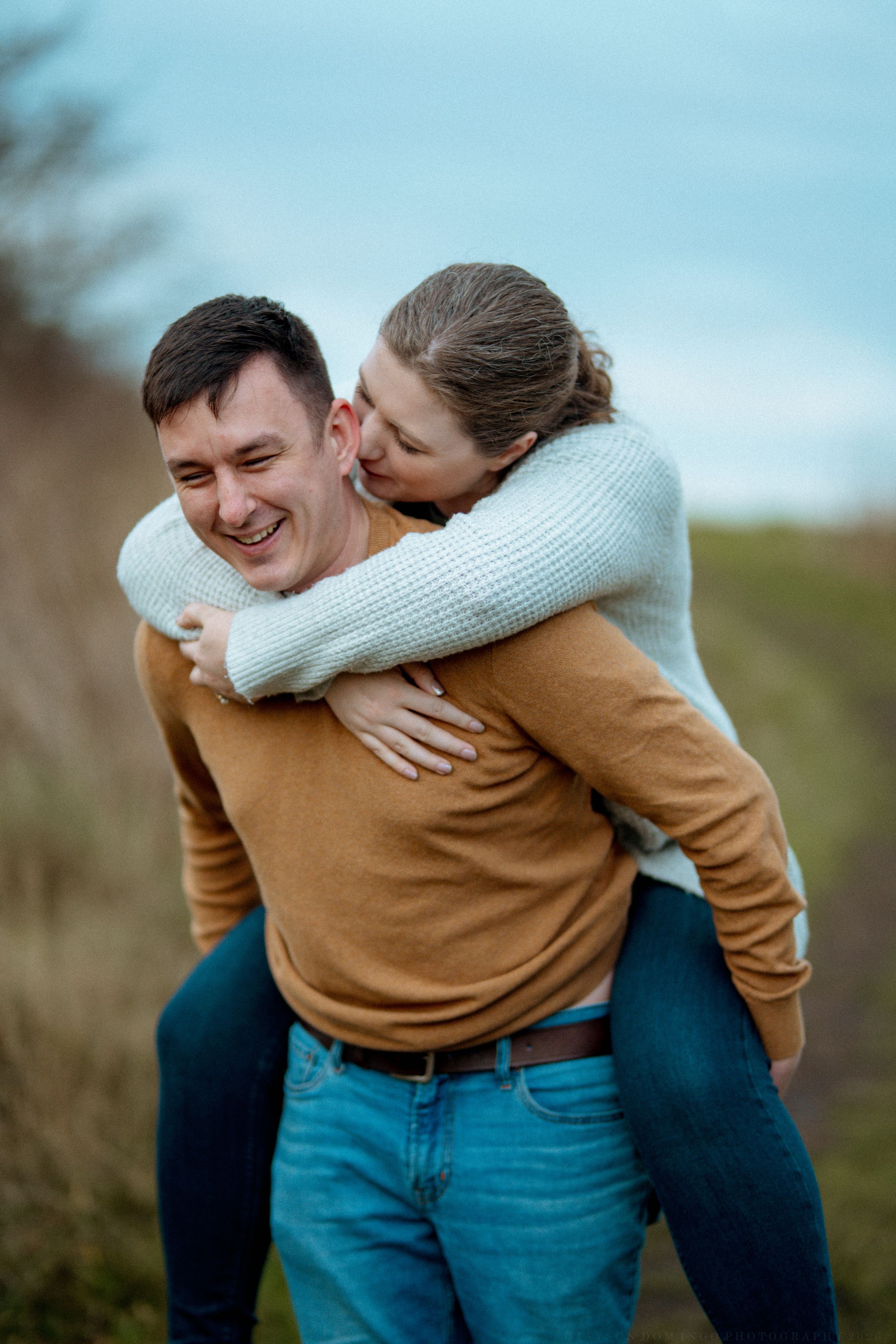 Two people embrace playfully outdoors with one wearing a camel sweater and the other in white.
