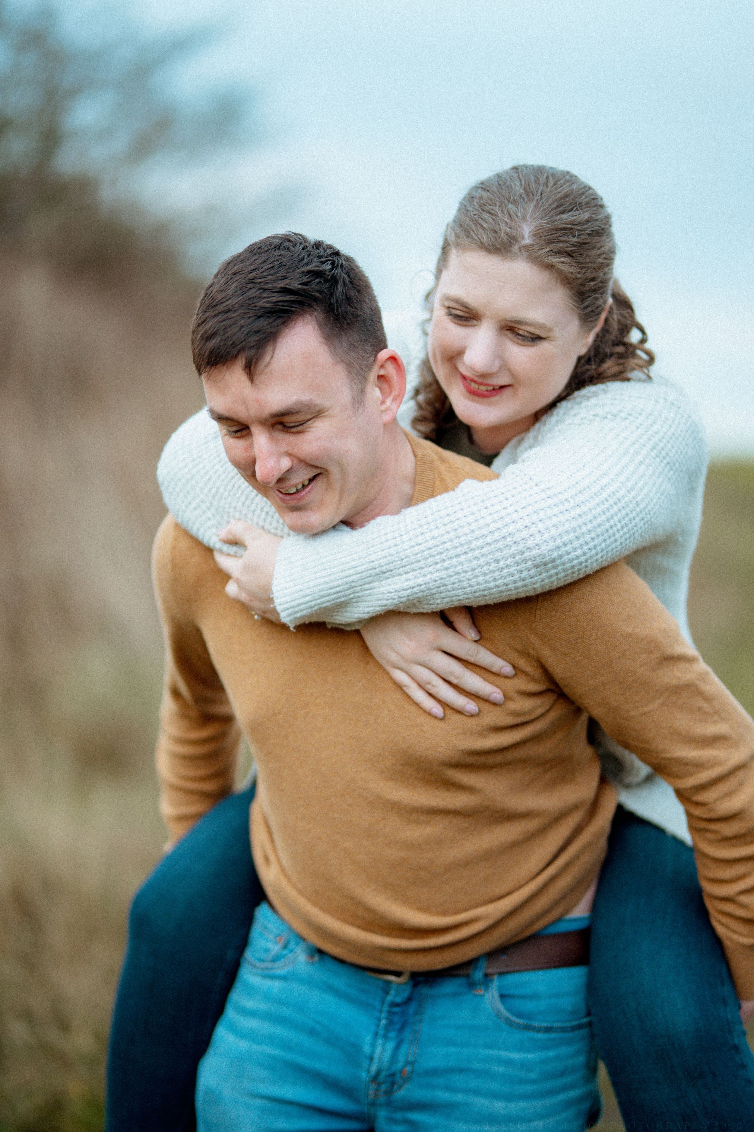 A joyful piggyback ride outdoors with a couple wearing casual sweaters on a cloudy day.