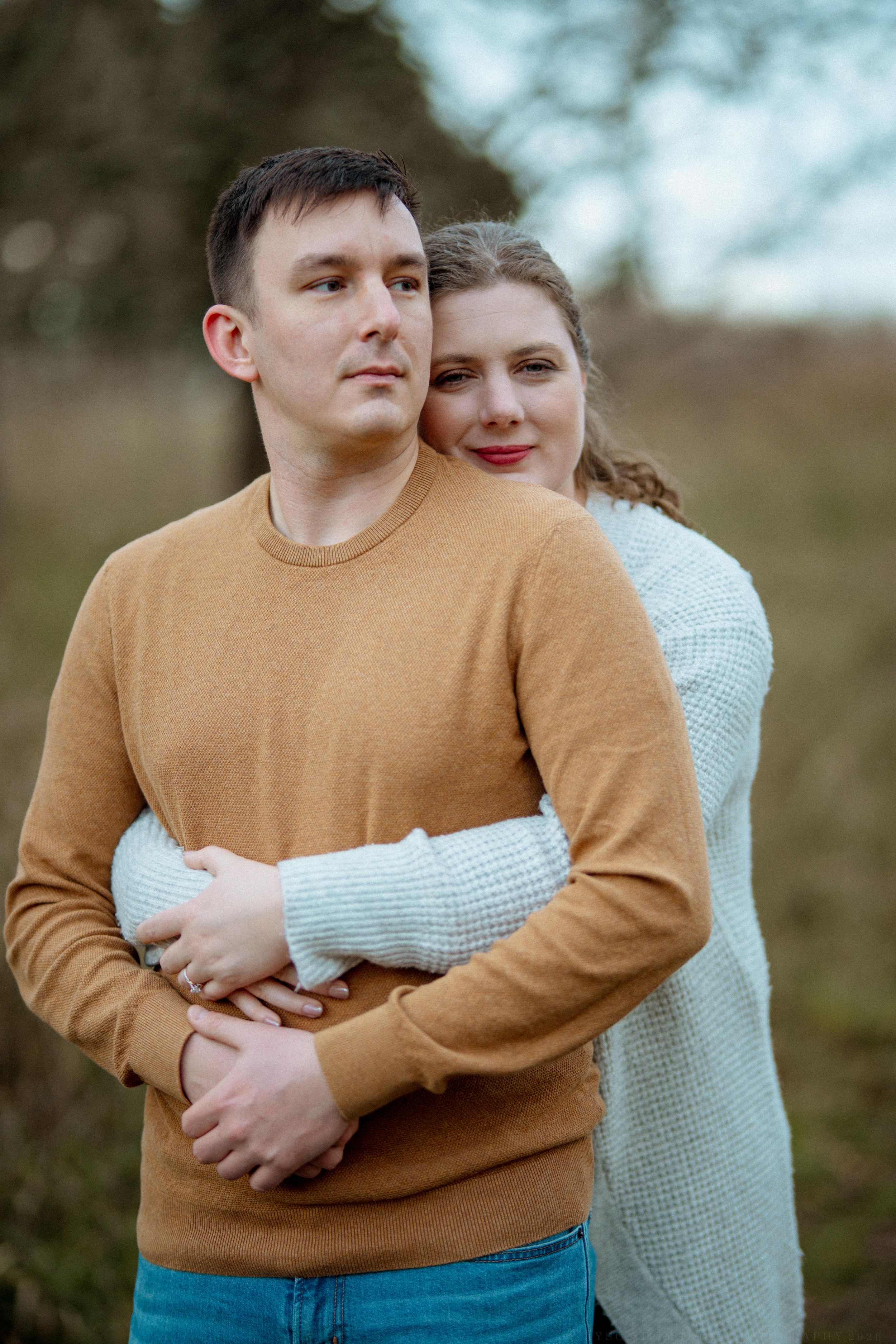 A romantic outdoor portrait of a couple hugging, with one wearing a tan sweater and the other in white.