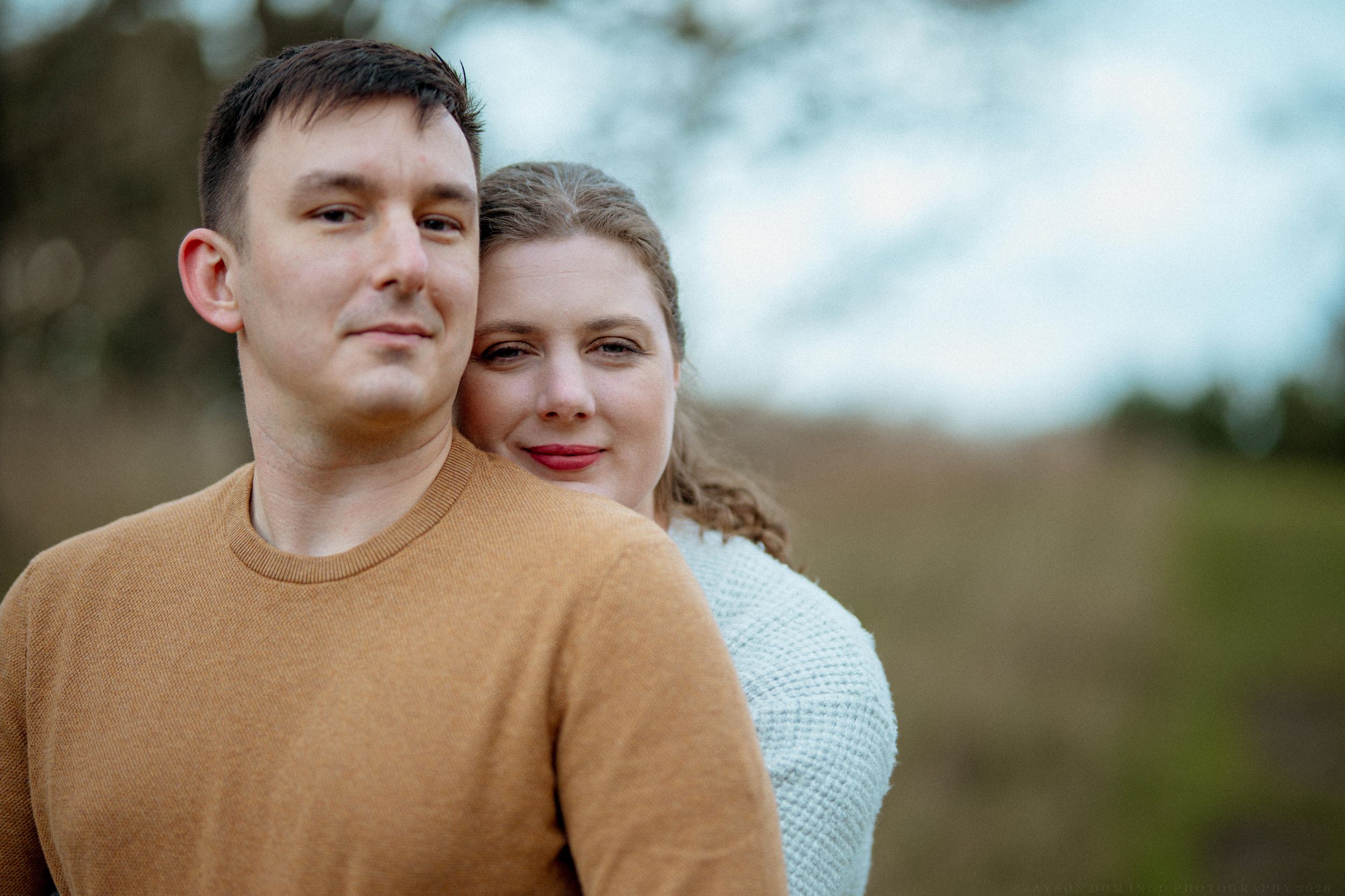 A couple in casual attire embraces outdoors during a golden hour photoshoot in a natural setting.