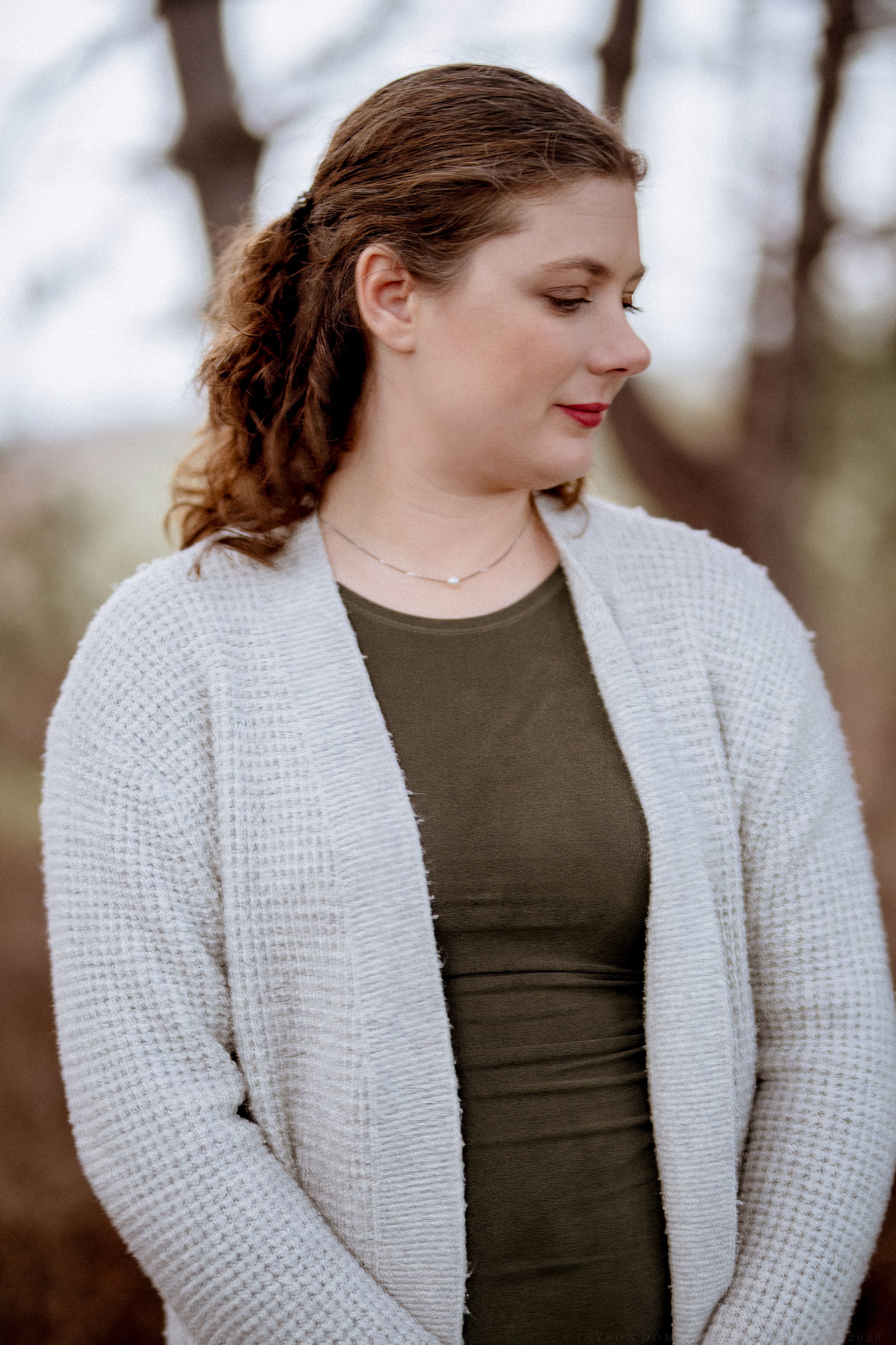 A woman in a white knit cardigan and olive top stands outdoors with a soft, contemplative expression.