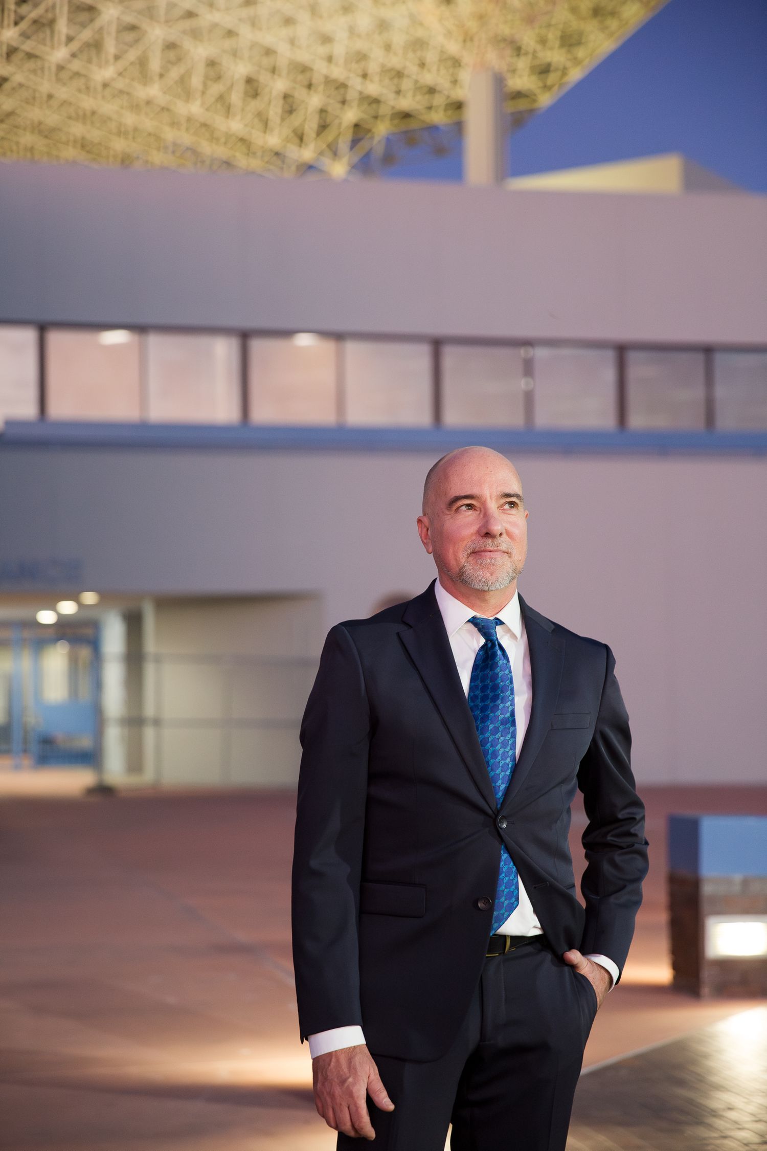 Professional in a black suit and blue tie stands confidently against modern architectural backdrop at dusk.