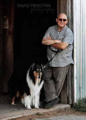 A person in sunglasses sits in a doorway with a black and white Shetland Sheepdog in a rustic barn setting.