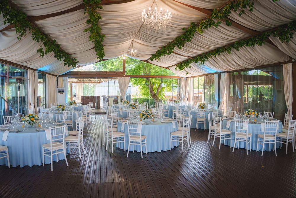 Elegant wedding reception venue with draped ceiling, crystal chandeliers, blue linens and white chairs under a white tent.