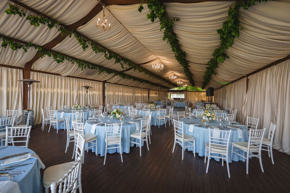 Elegant wedding reception venue with draped white fabric ceiling, green garlands, and white Chiavari chairs arranged in rows.