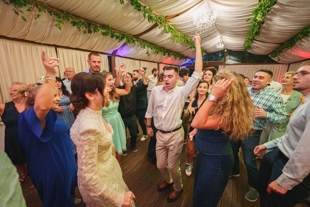 Wedding guests dance and celebrate under a white tent decorated with green foliage garlands during a lively reception.