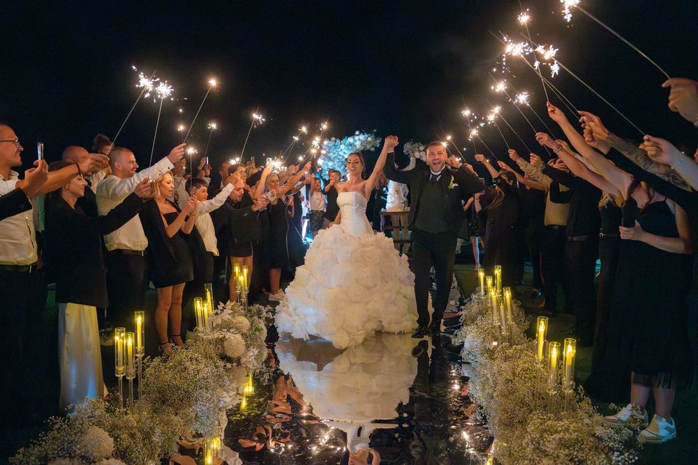 Wedding guests wave sparklers in a circle around a couple in white during an outdoor nighttime celebration.