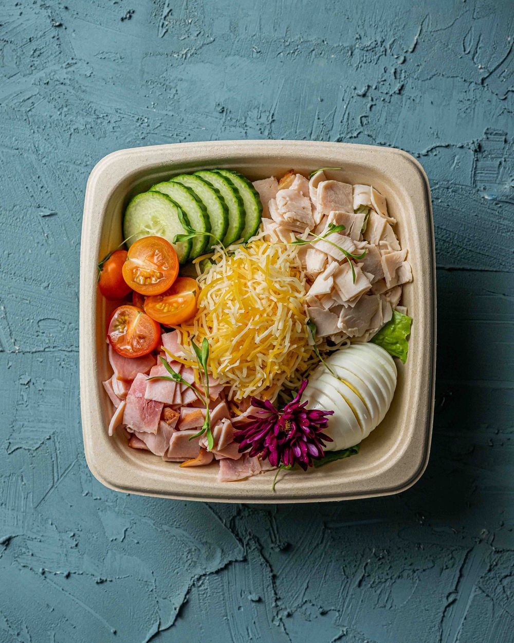 Fresh salad with shredded cheese, cucumber slices, tomatoes and ham served in a takeout container on blue surface.
