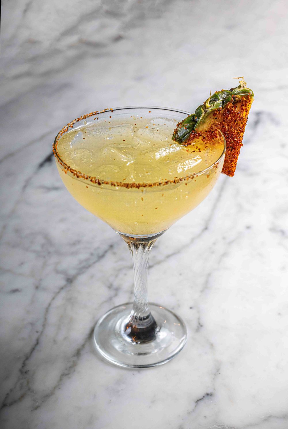 A golden margarita cocktail garnished with chili powder rim and fresh herbs served in a coupe glass on marble surface.