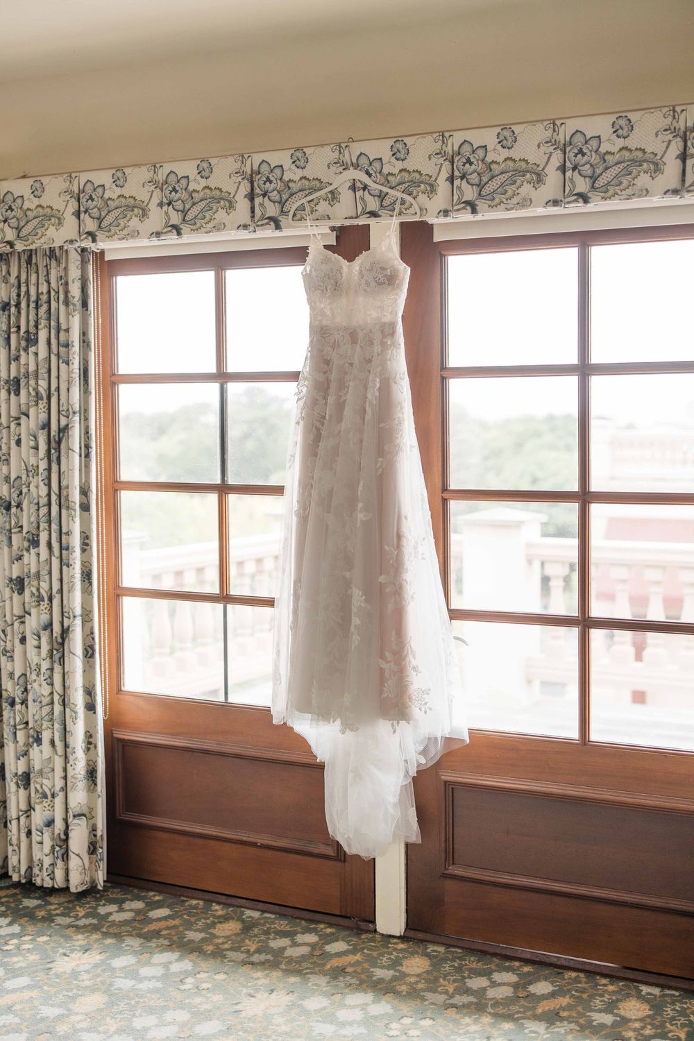 A white wedding dress hangs in front of large windows with floral curtains in a sunlit room.