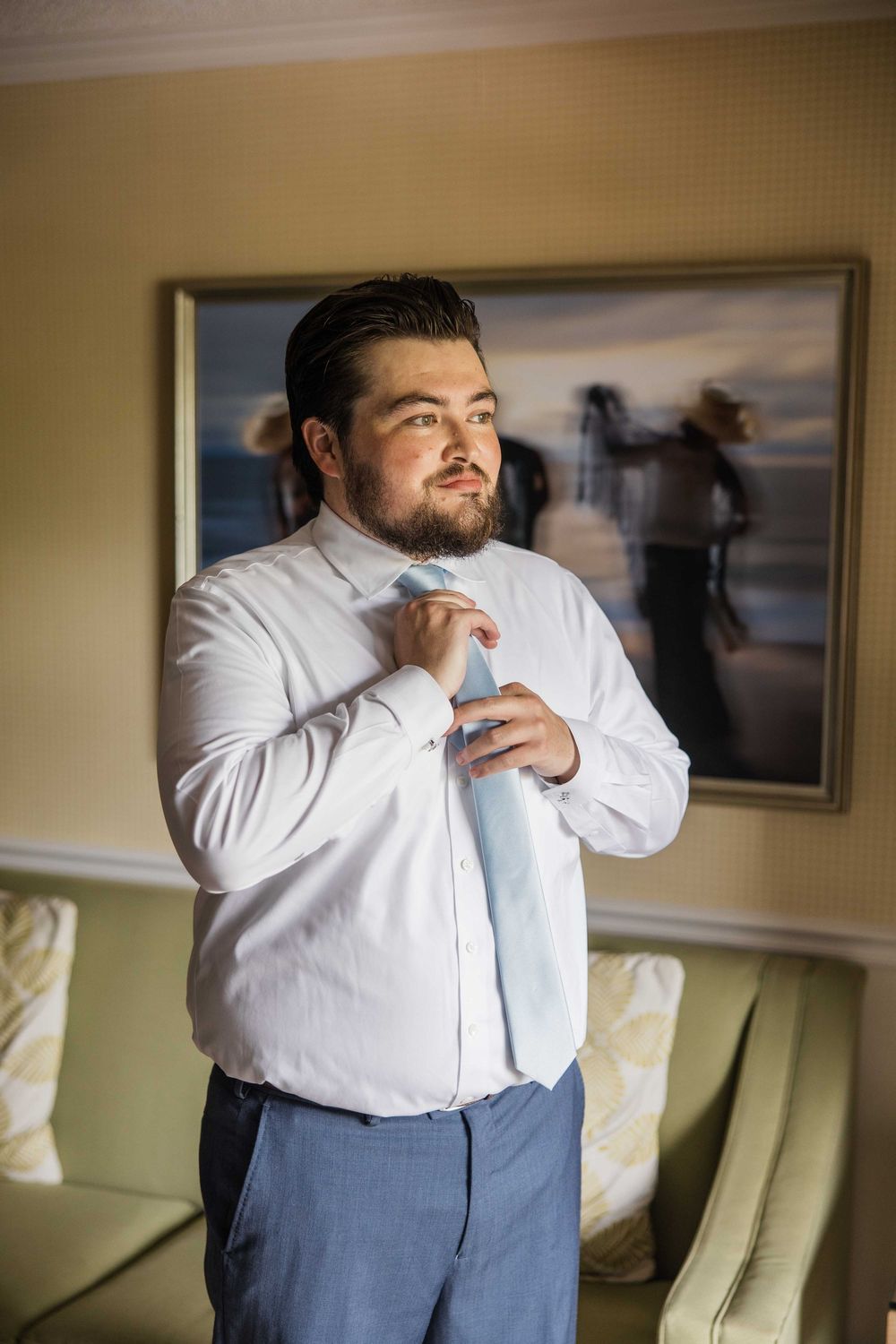 A person in a white dress shirt and blue tie adjusts their clothing while getting ready in a room.
