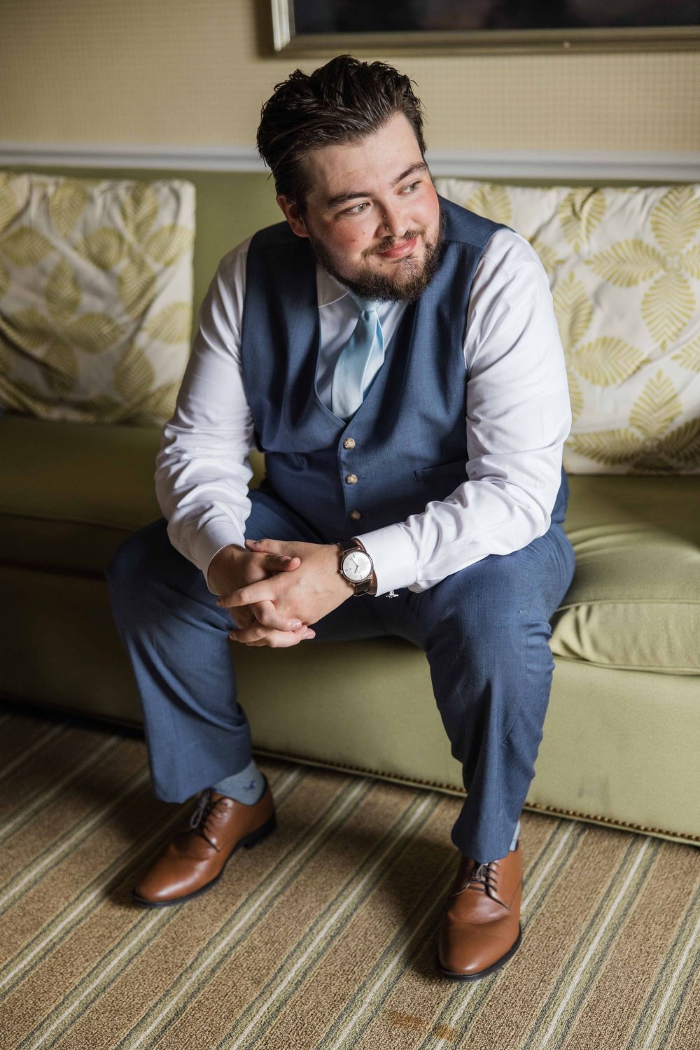 A person in formal attire with a blue vest sits on a green couch, wearing brown leather shoes and a white dress shirt.