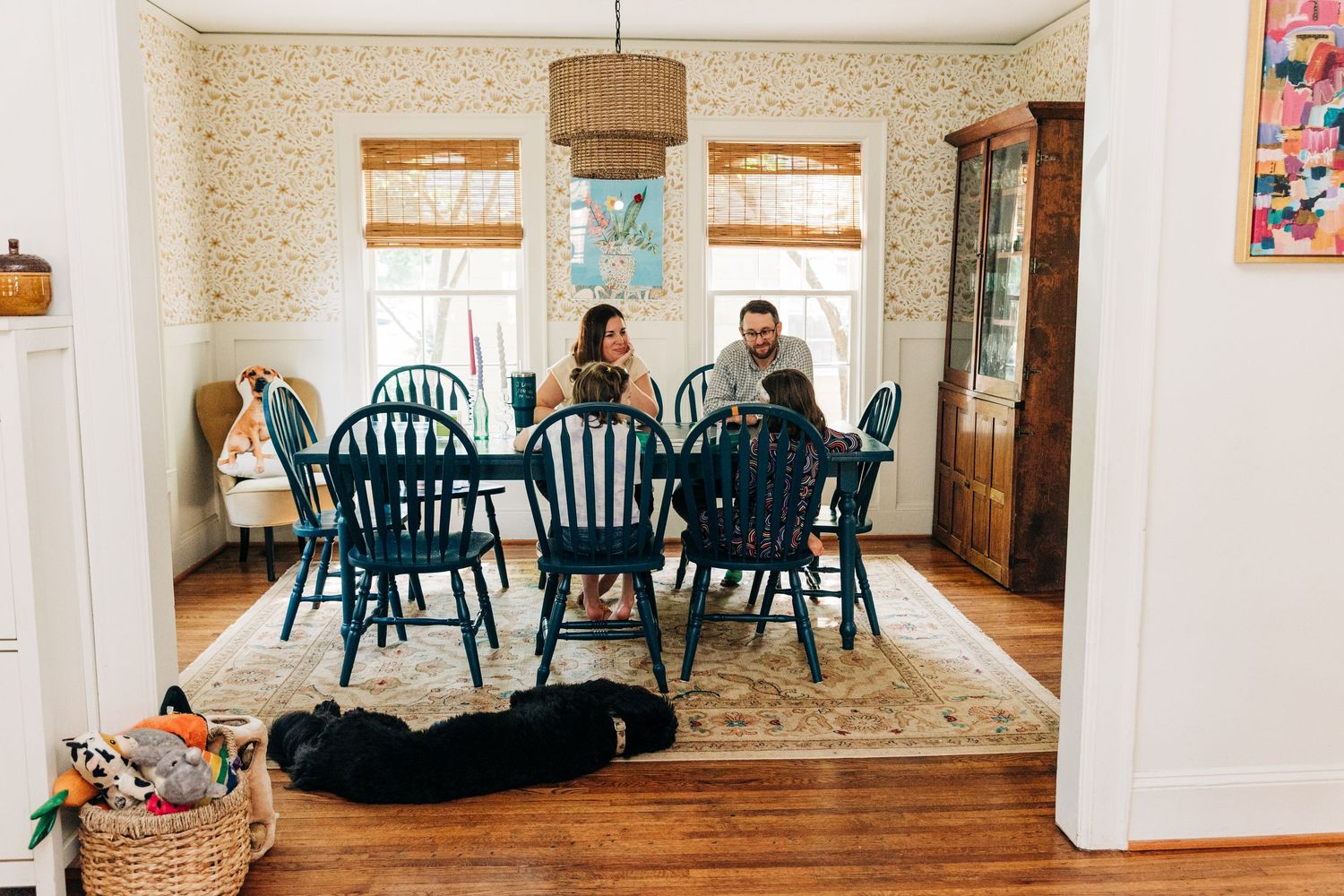 A cozy dining room with teal blue chairs around a wooden table and a black dog resting on the floor. A Charlotte family plays cards.