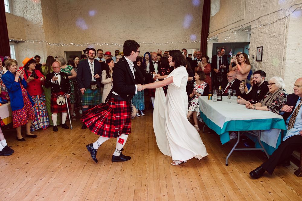 A couple dances at a wedding reception with the groom wearing a traditional Scottish kilt and the bride in white.