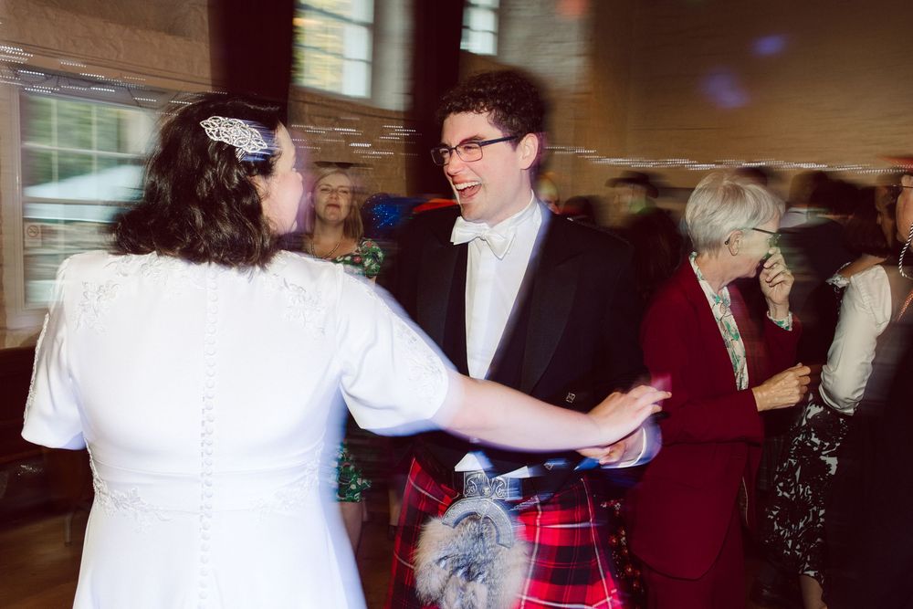 A couple dances together at a wedding reception, with the groom wearing a kilt and bowtie while spinning his partner.