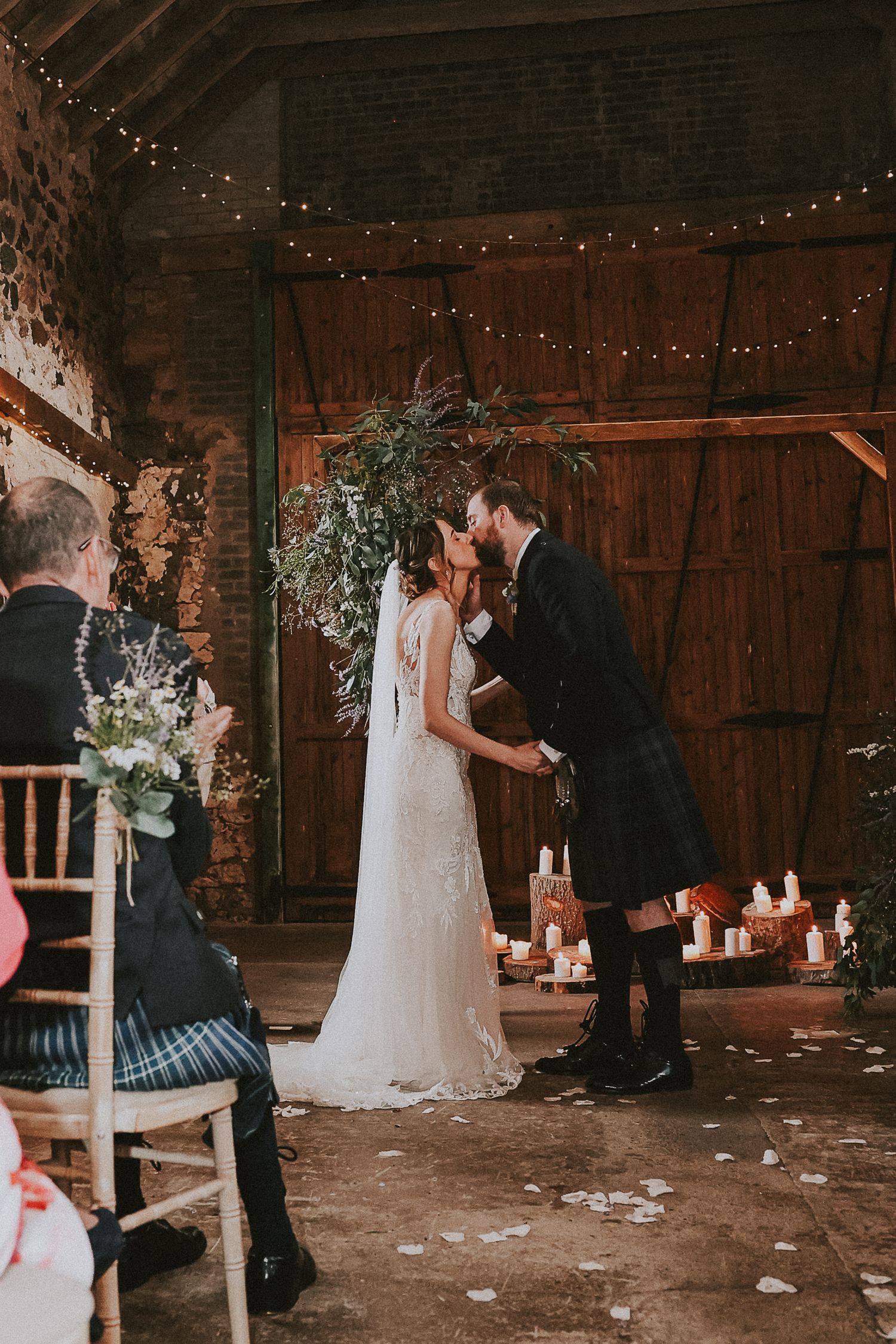 Intimate wedding ceremony in rustic barn venue with twinkling lights and greenery.