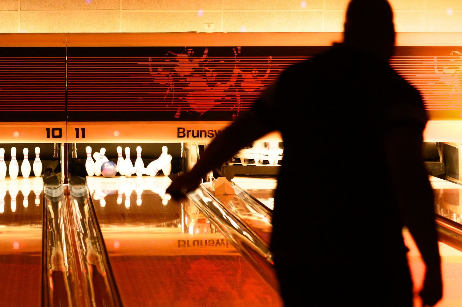 A silhouetted figure stands at a bowling alley lane with glowing pins and warm orange lighting in the background.
