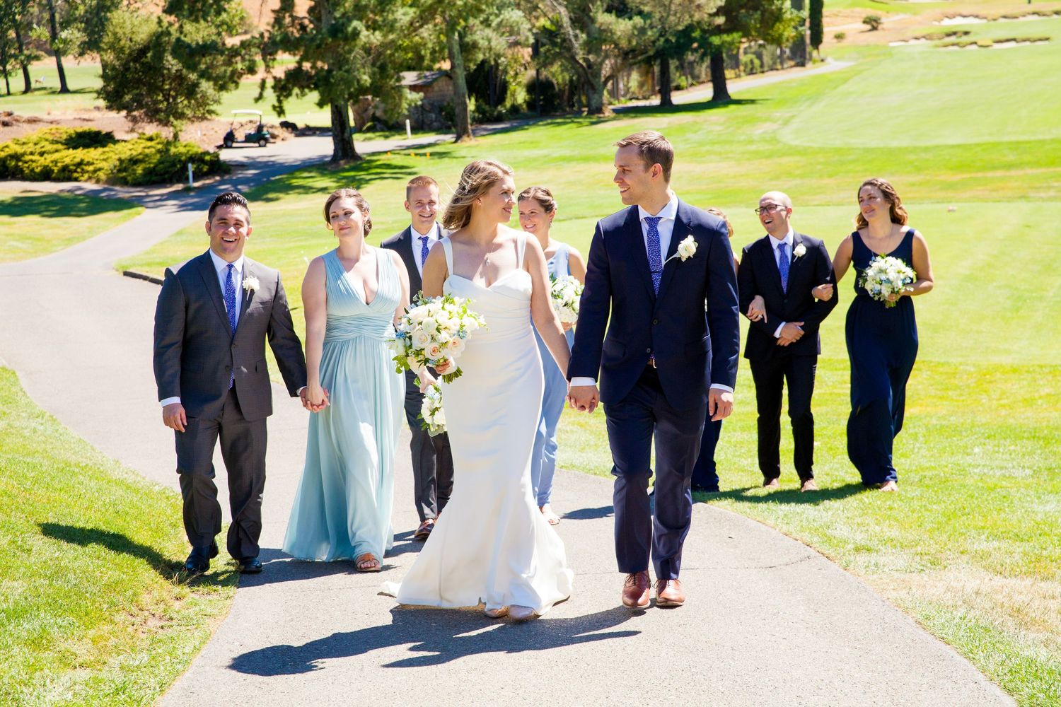 Wedding party members walk together on a sunny day across a lush green lawn during an outdoor celebration.