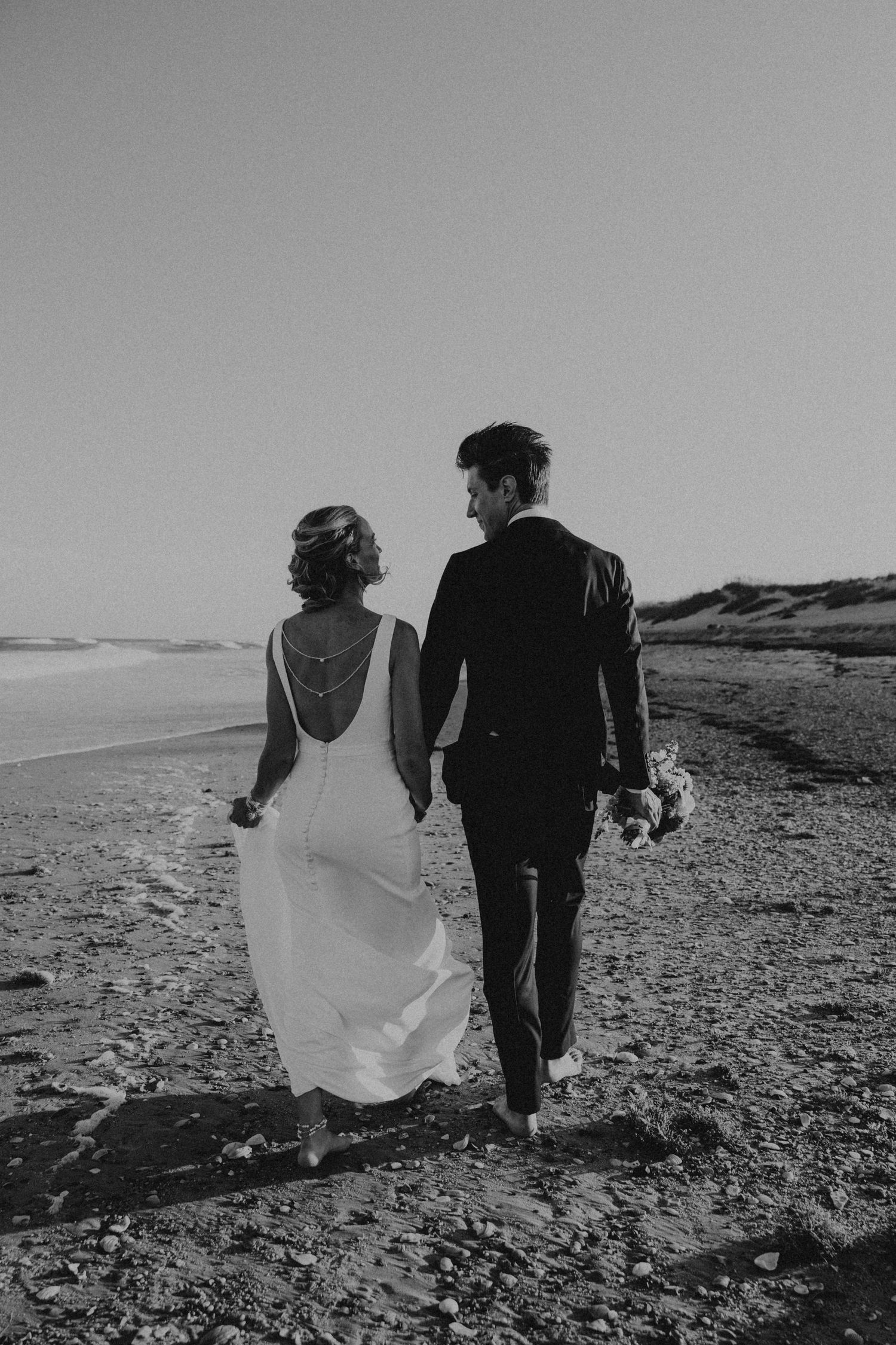 A couple walks along a rocky beach shoreline in black and white as the bride's dress flows in the wind.