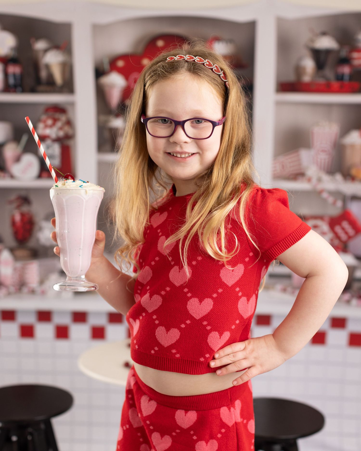 A milkshake in a tall glass with red heart decor and retro diner shelving visible in the background.