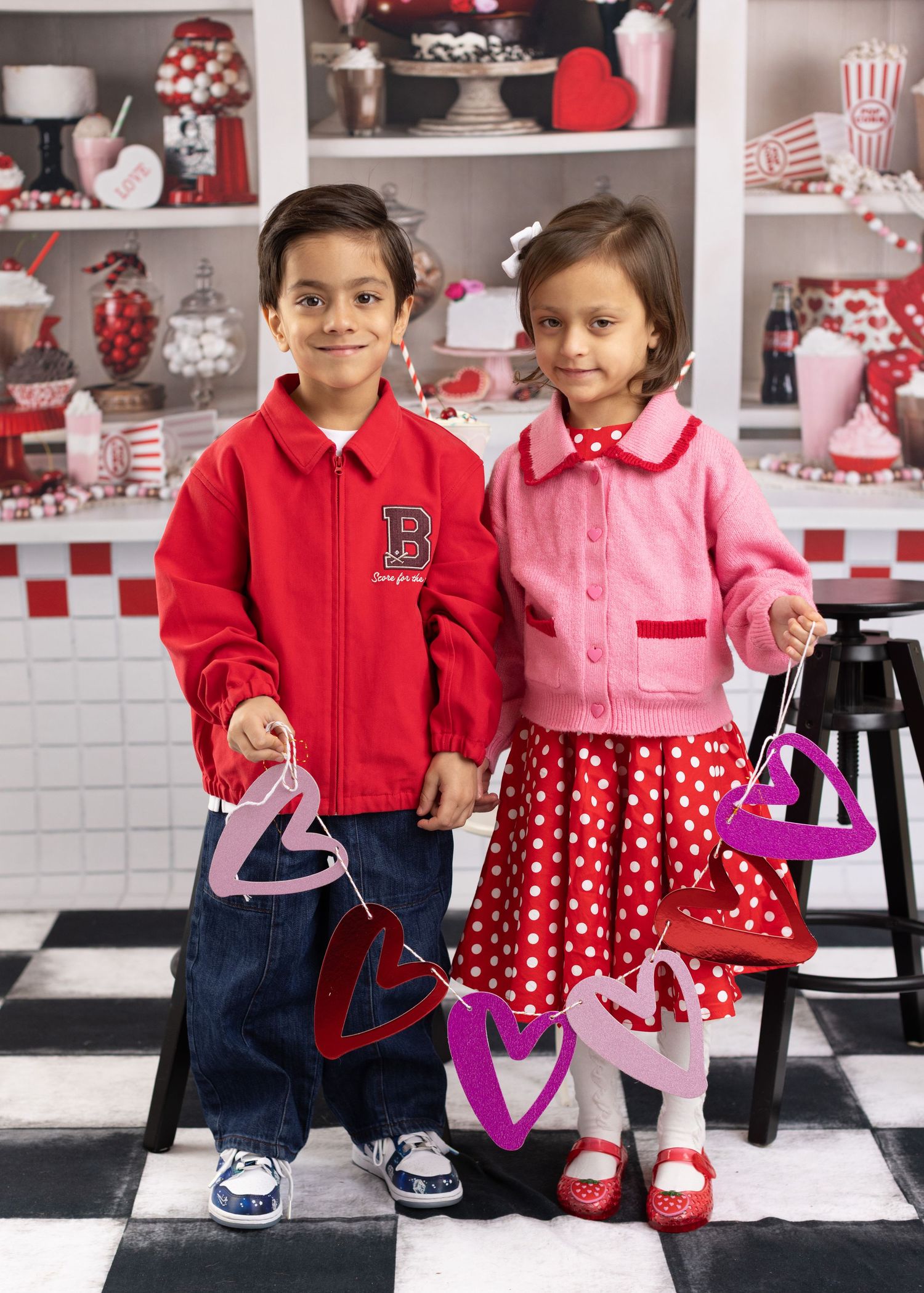 Two children in coordinating red and pink outfits pose together in front of a retro diner-style backdrop with polka dot decor.