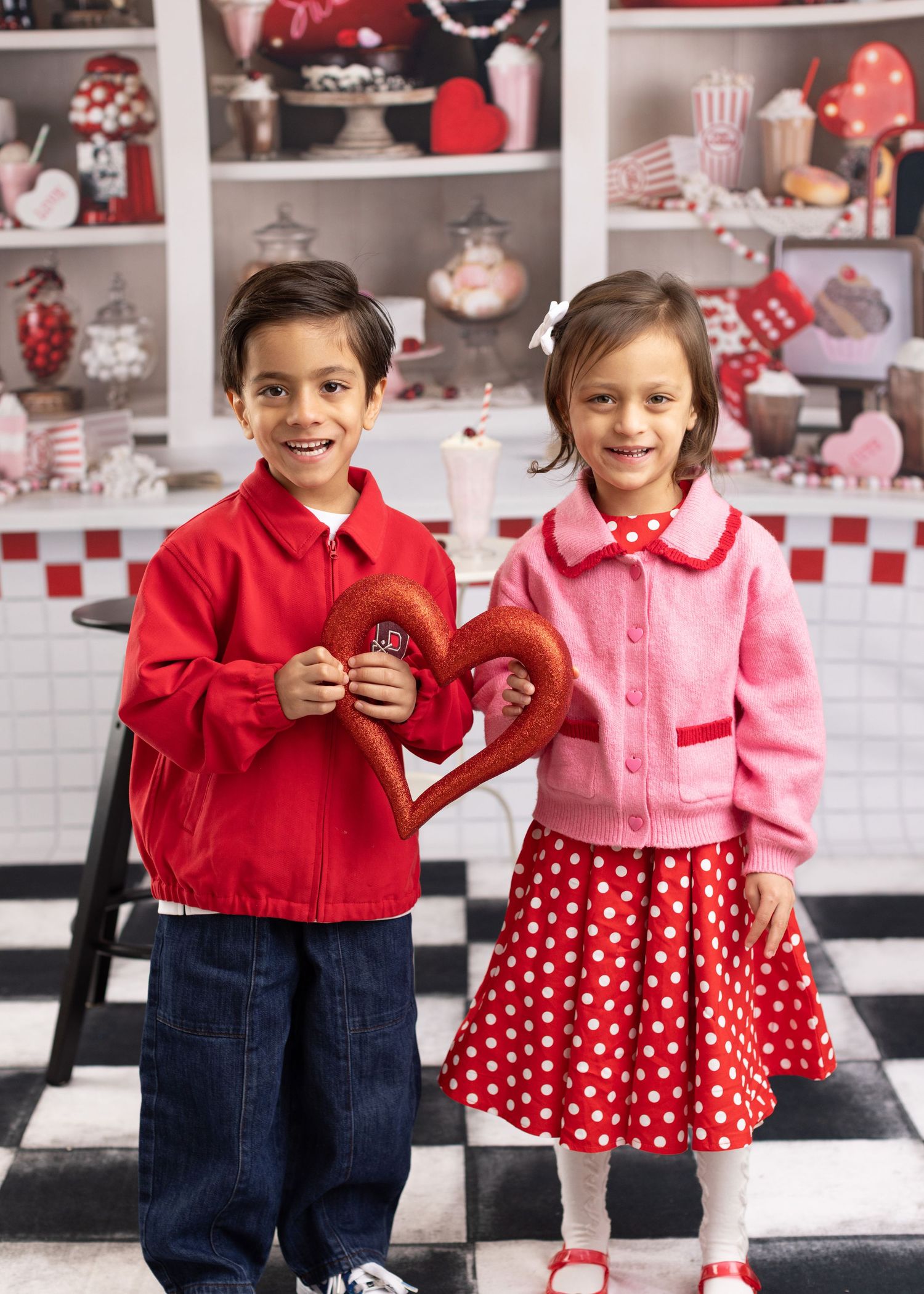 Two children in retro-style red and pink outfits pose together in a diner-themed setting with checkerboard floors.
