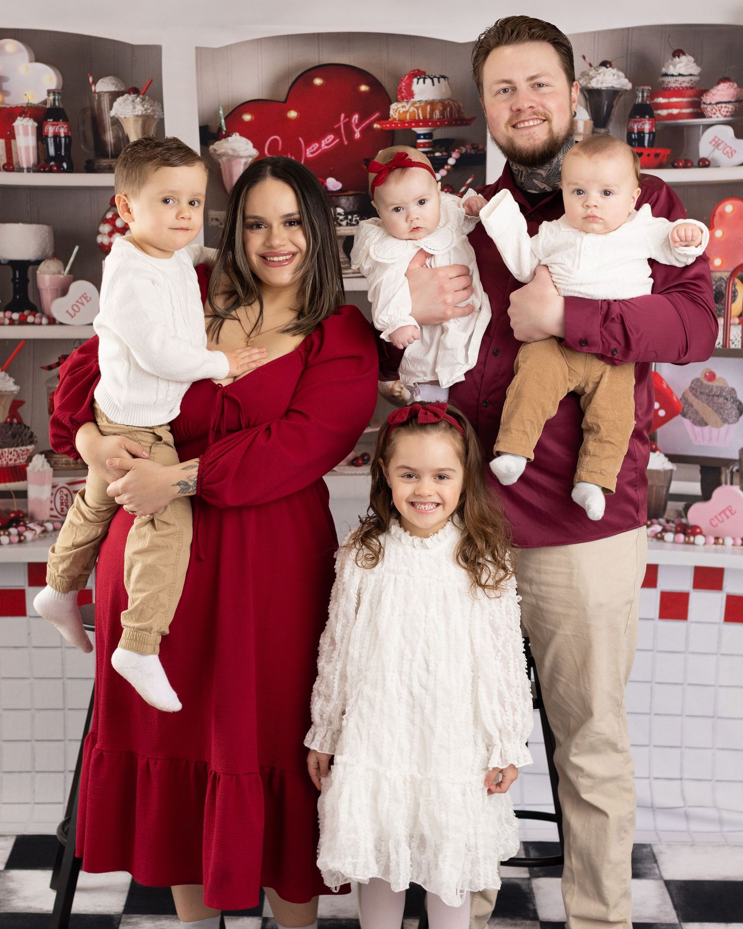 Family poses together in coordinated burgundy and white holiday outfits against a white brick wall with festive decor.