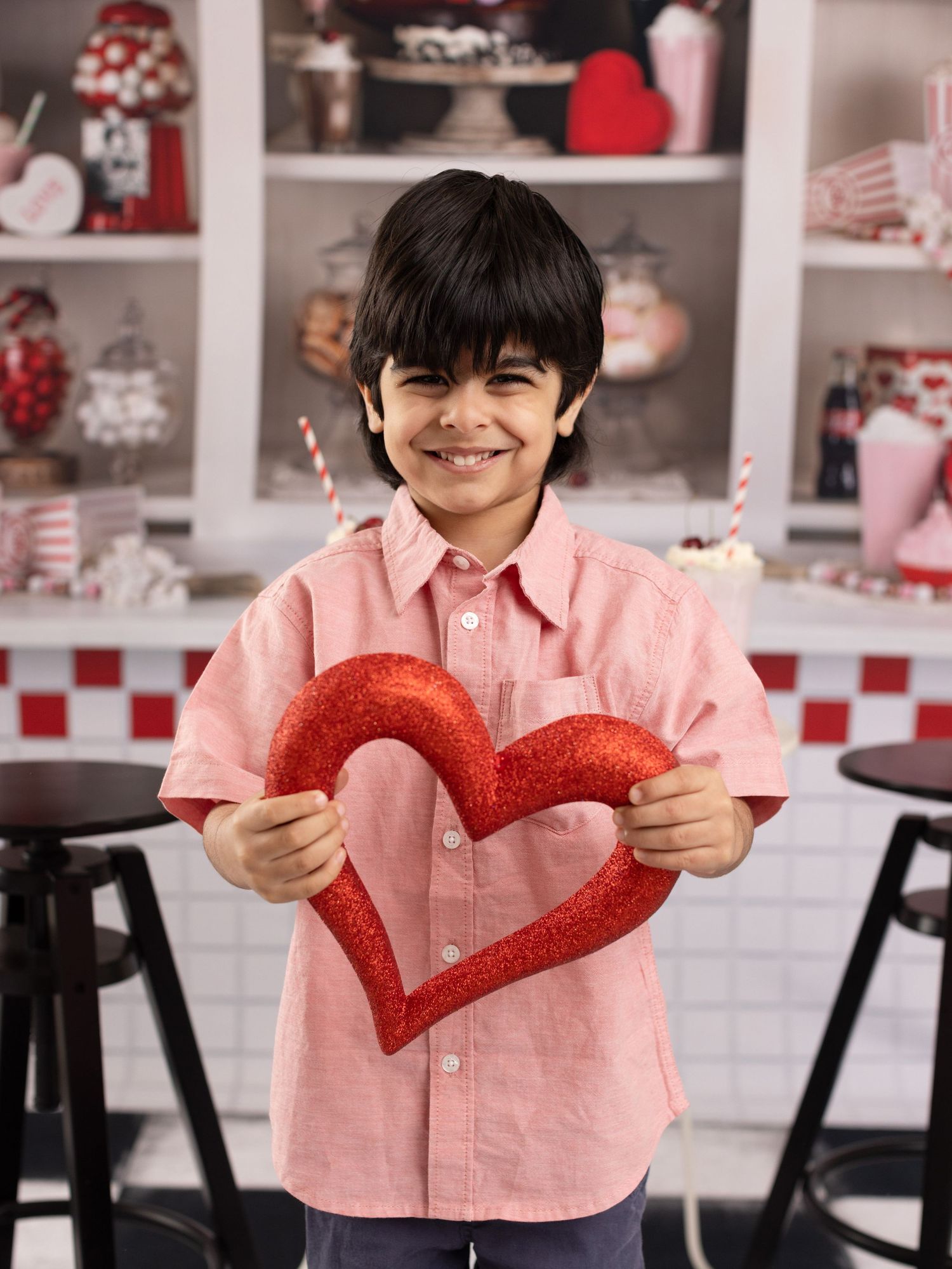 A young person in a pink shirt holds up a glittery red heart decoration in a kitchen setting with Valentine's decor.