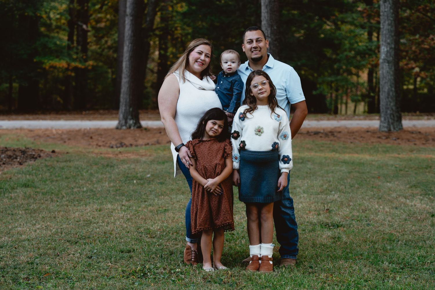 A family stands together outdoors in front of tall trees during an autumn photo session.