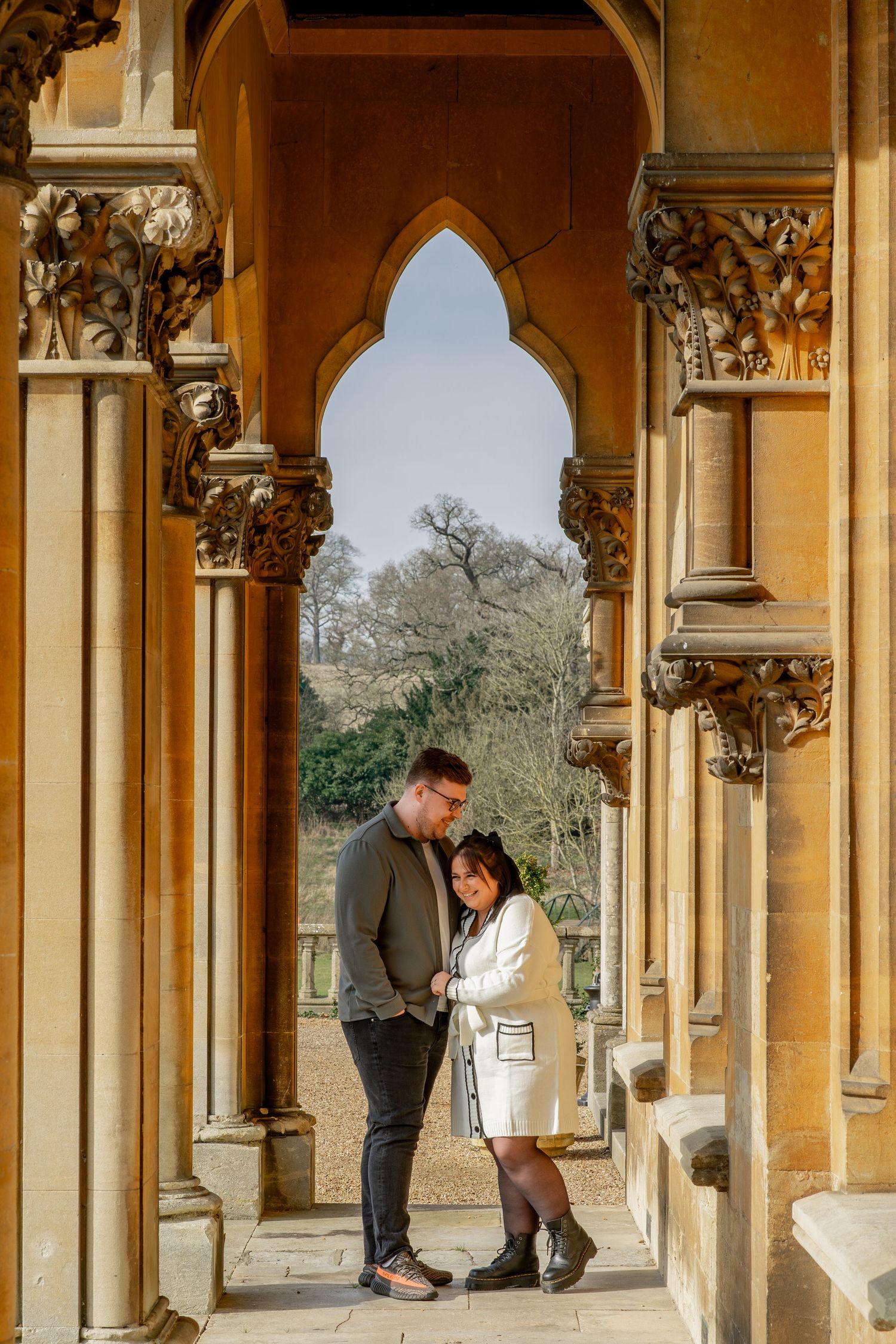 A couple shares an intimate moment under ornate Gothic stone archways at a historic building with golden columns.