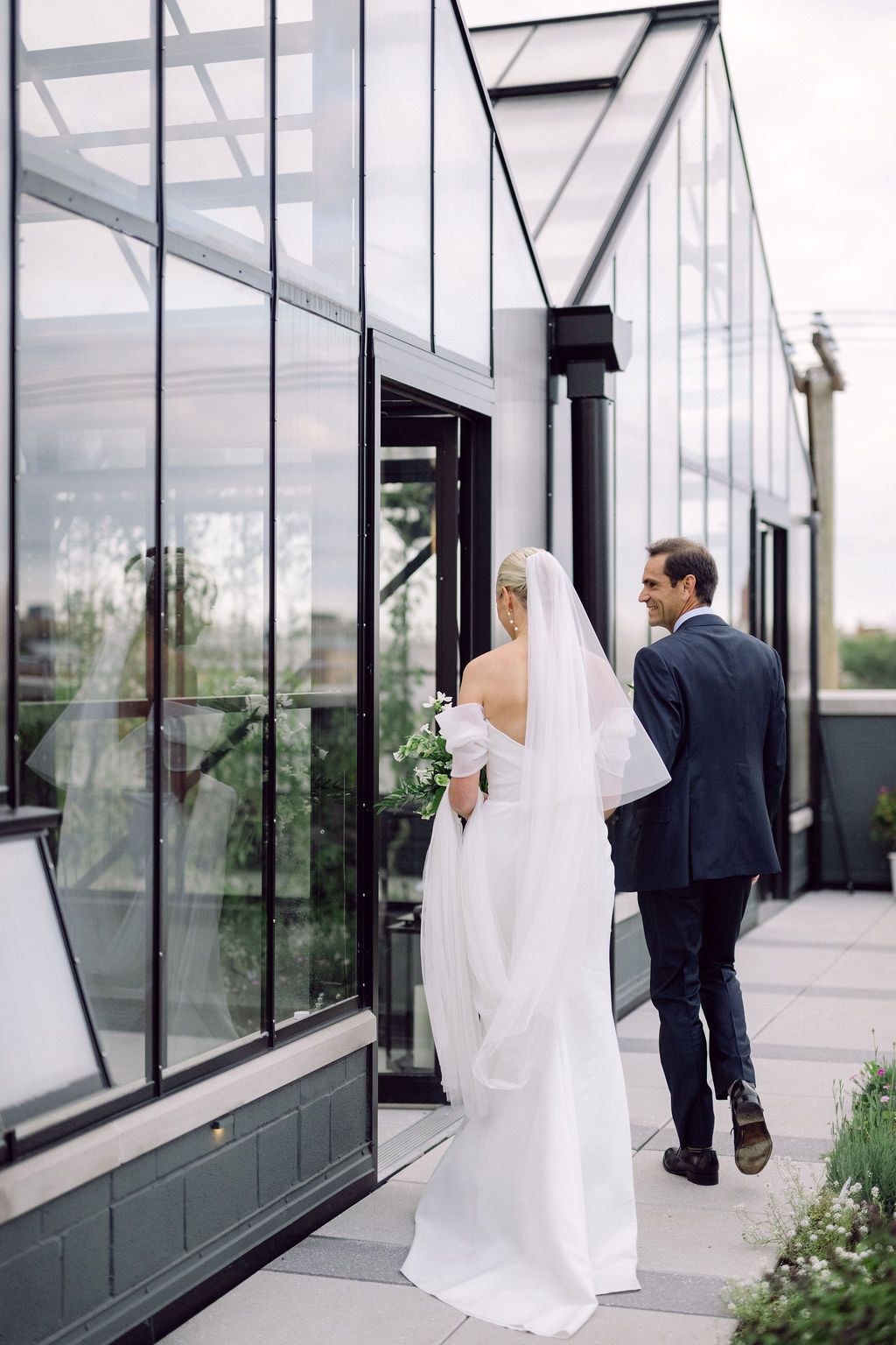 A black and white wedding ceremony photo with a couple exchanging vows under a floral arch against a brick wall.