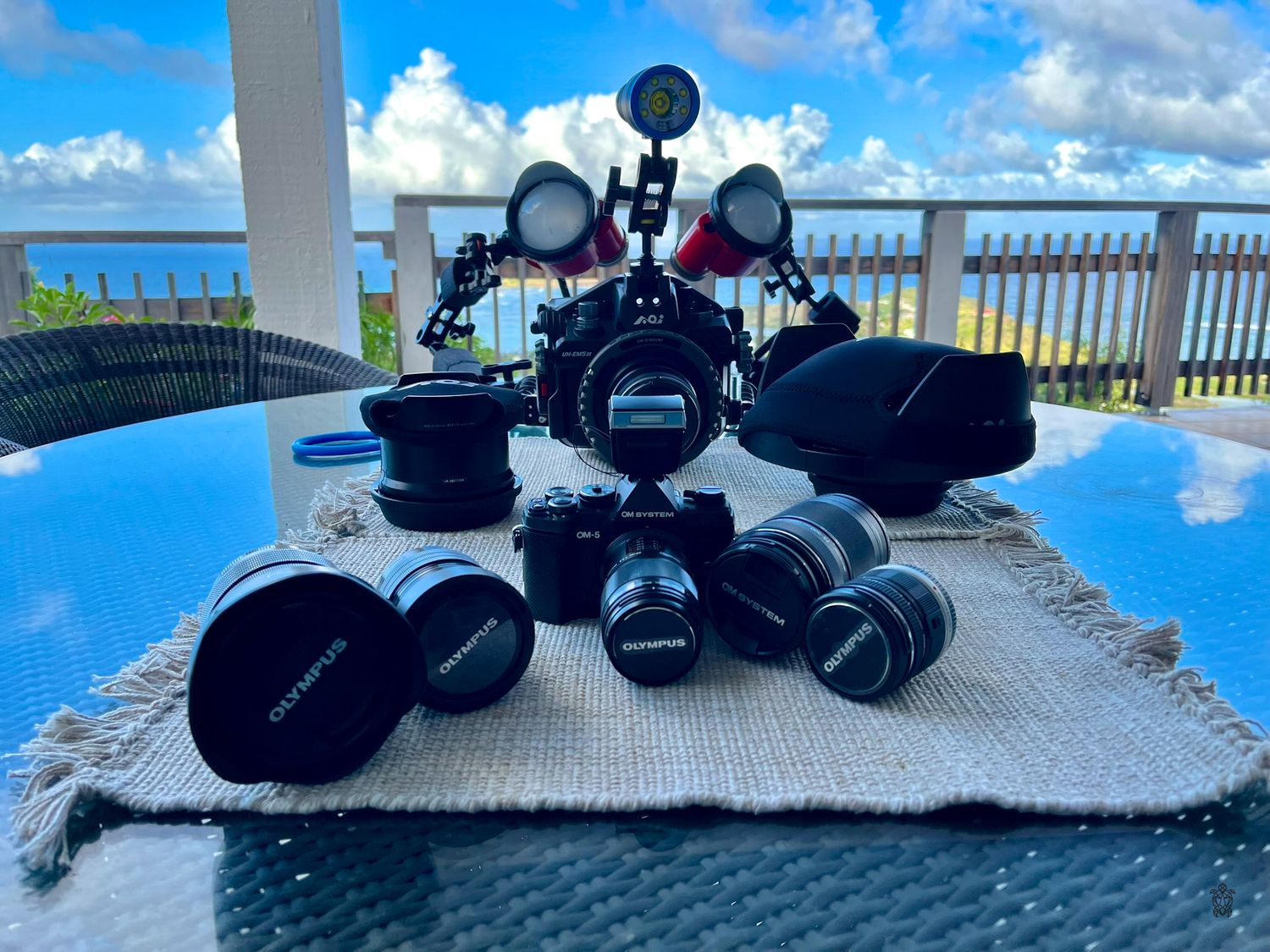 Camera equipment and lenses displayed on blue table overlooking tropical ocean view with bright blue sky and clouds.