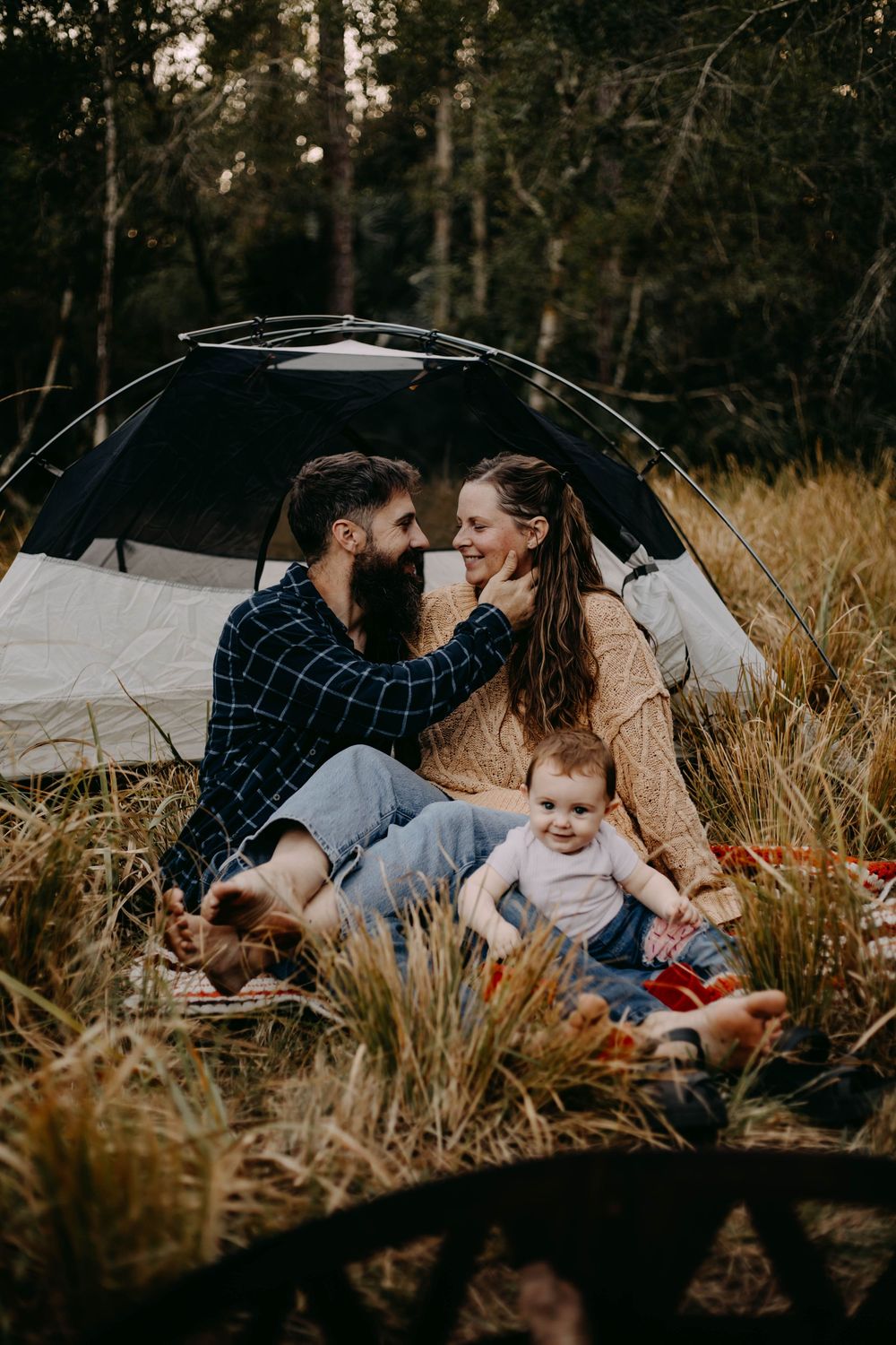 A family shares tender moments together while sitting in tall grass during an outdoor photoshoot at sunset.