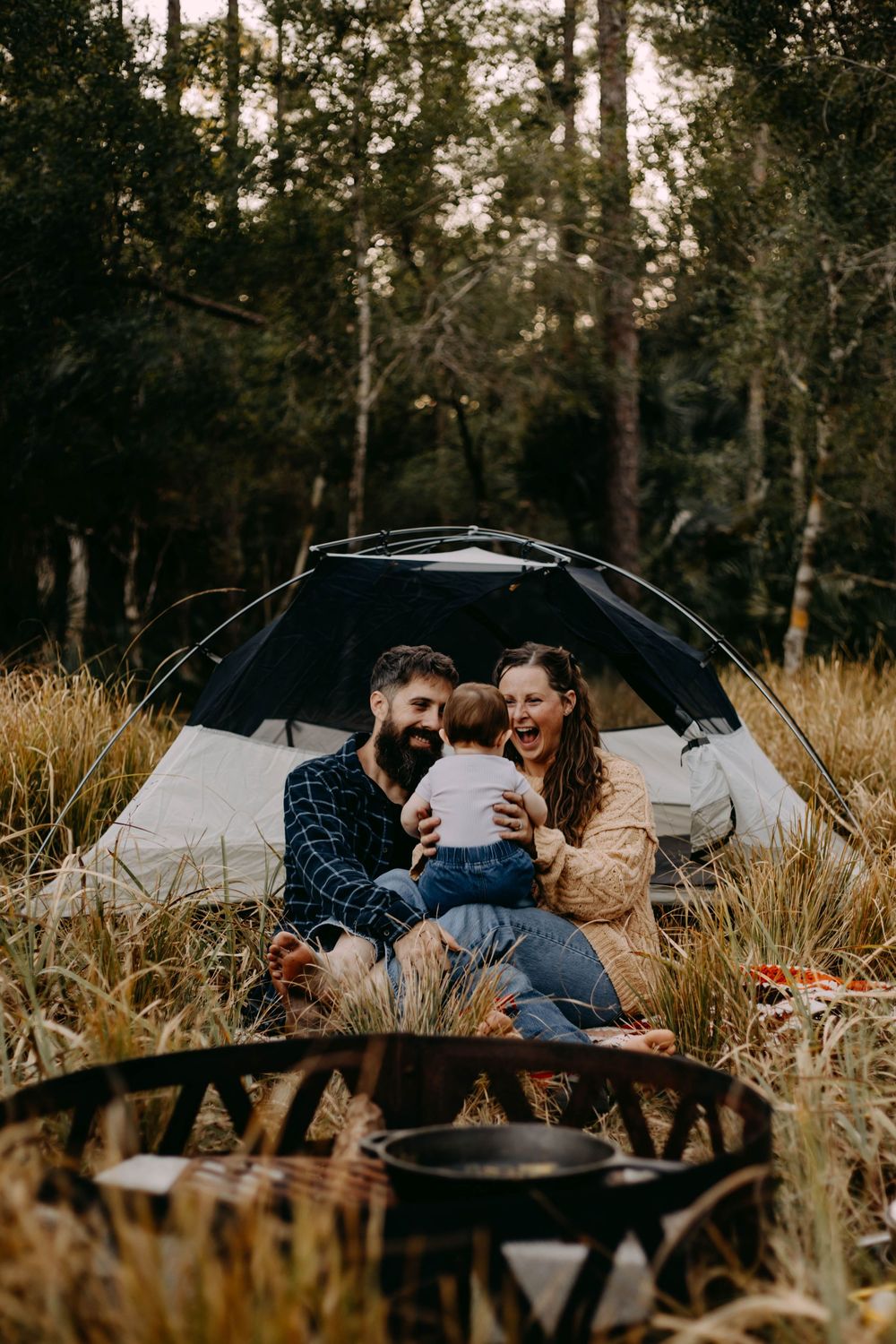 A couple relaxes together in a romantic outdoor setting surrounded by tall grass and trees at sunset.