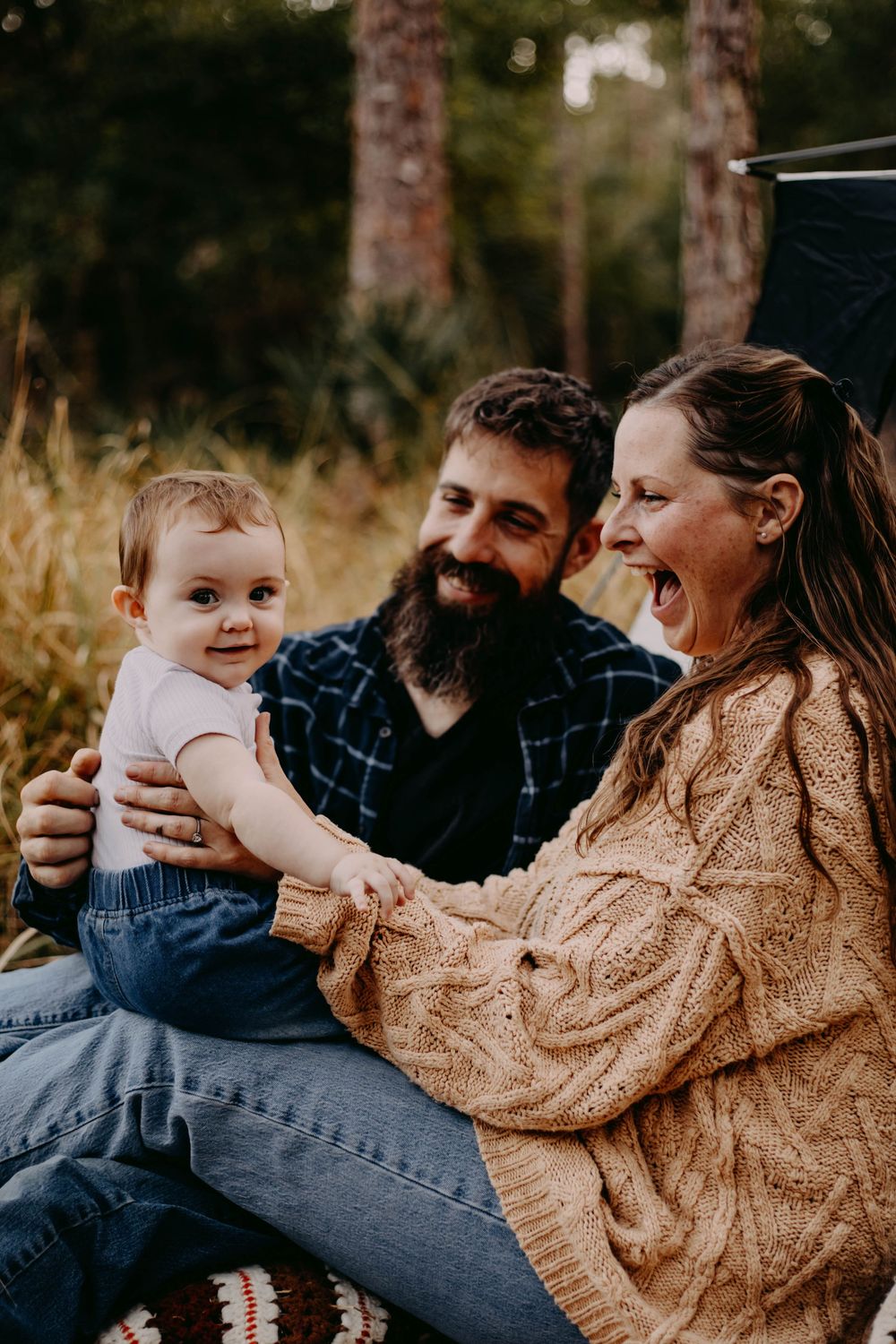 A family sits together in tall grass during golden hour wearing cozy sweaters and casual attire.