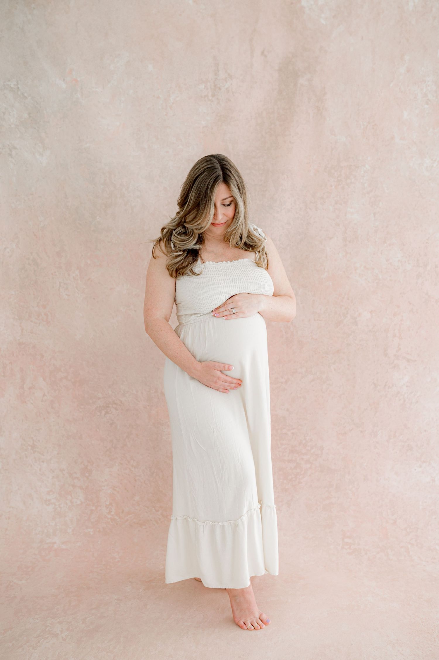 A pregnant woman in white dress against pink backdrop in an elegant pose showcasing the ​best time to do ​a maternity shoot.